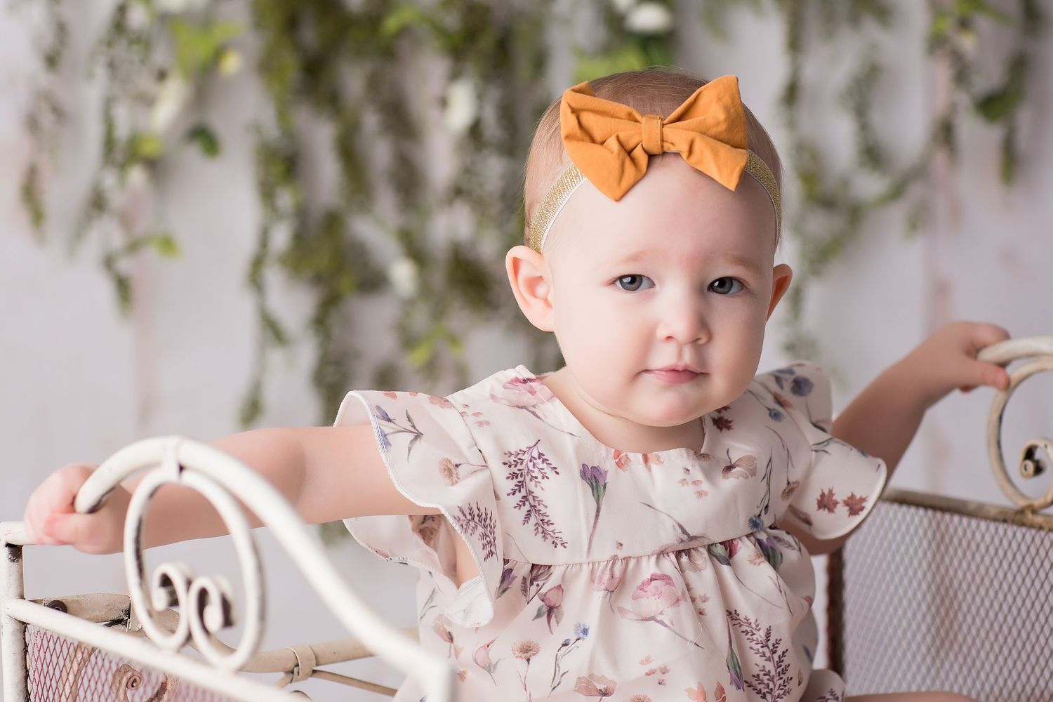 Baby wearing floral dress sits on white blanket with pink decorations in a studio setting.