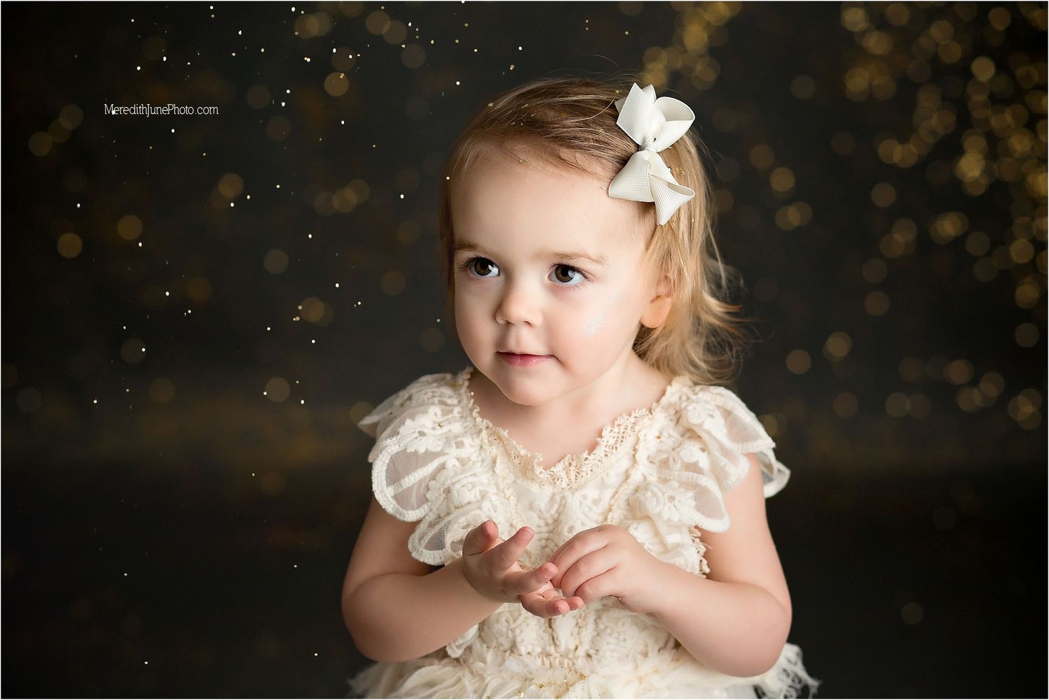 A child in a cream lace dress poses against a dark backdrop with floating gold glitter.