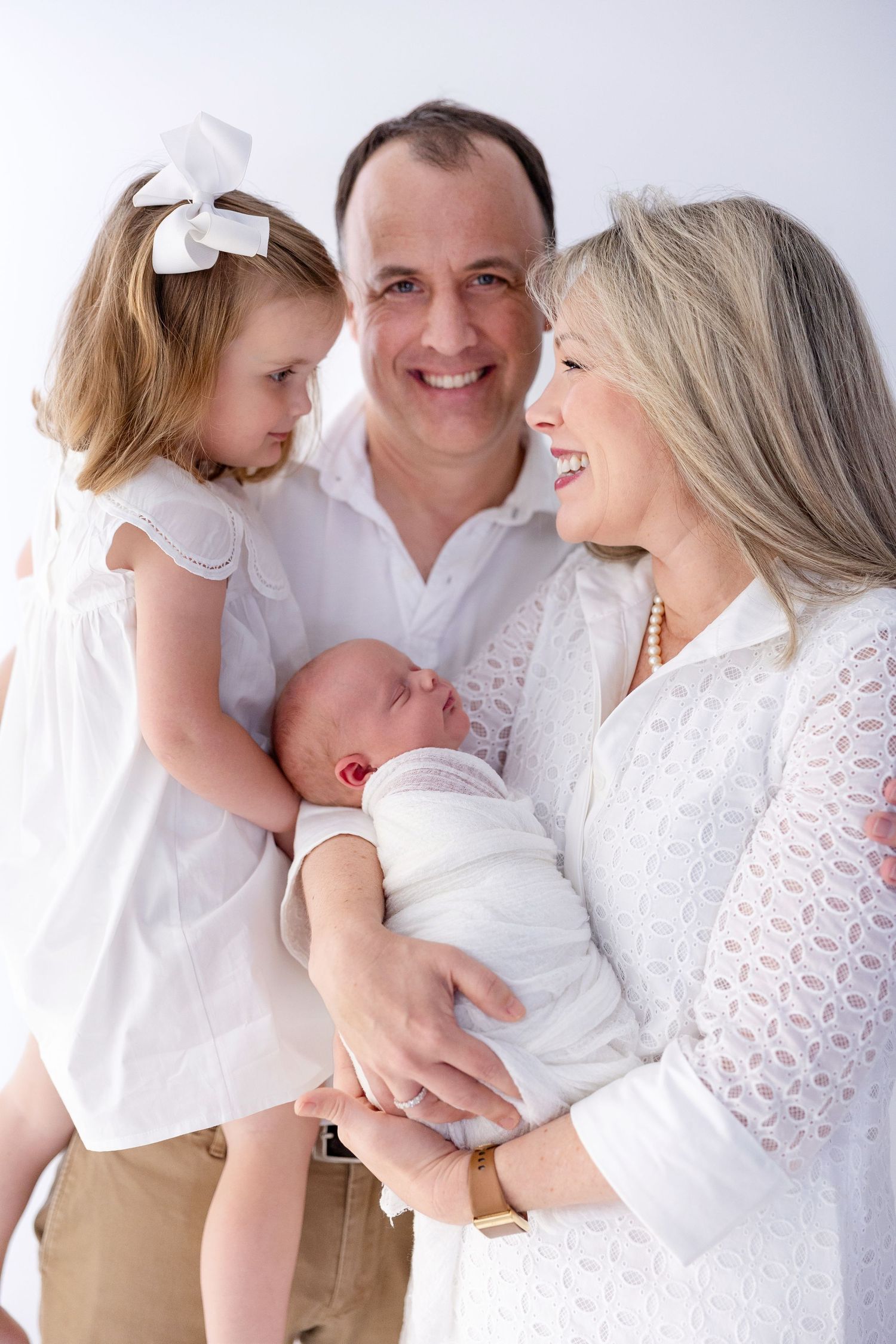 Grandparents smiling and holding newborn baby with toddler girl in white bow, family portrait.