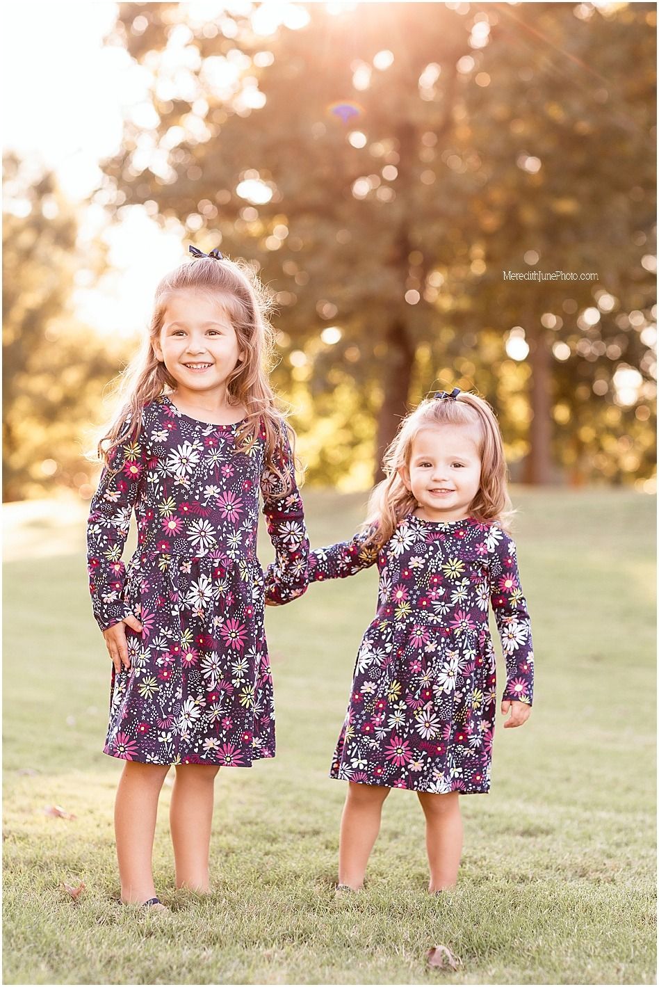 Two children in matching floral dresses playing together on grassy field during golden hour sunset.