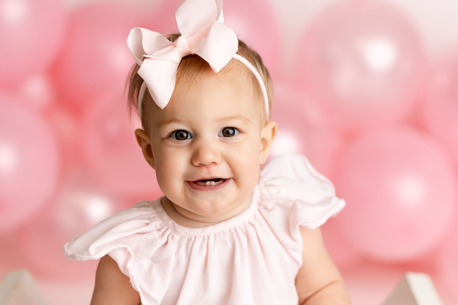 Adorable baby in white dress smiles against pink bokeh background with floral hair bow accent.