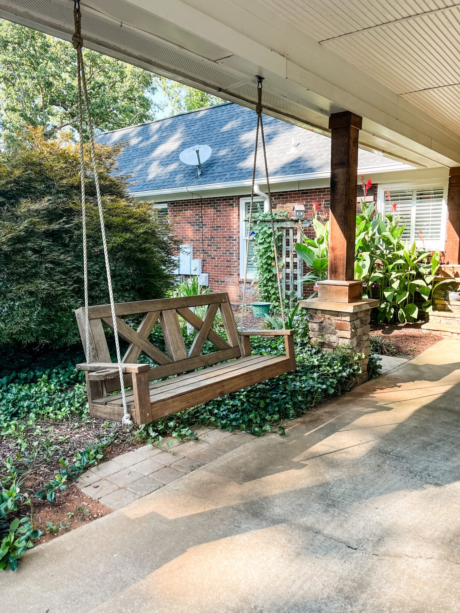 A rustic wooden porch swing hangs from chains on a covered patio surrounded by lush greenery and brick walls.