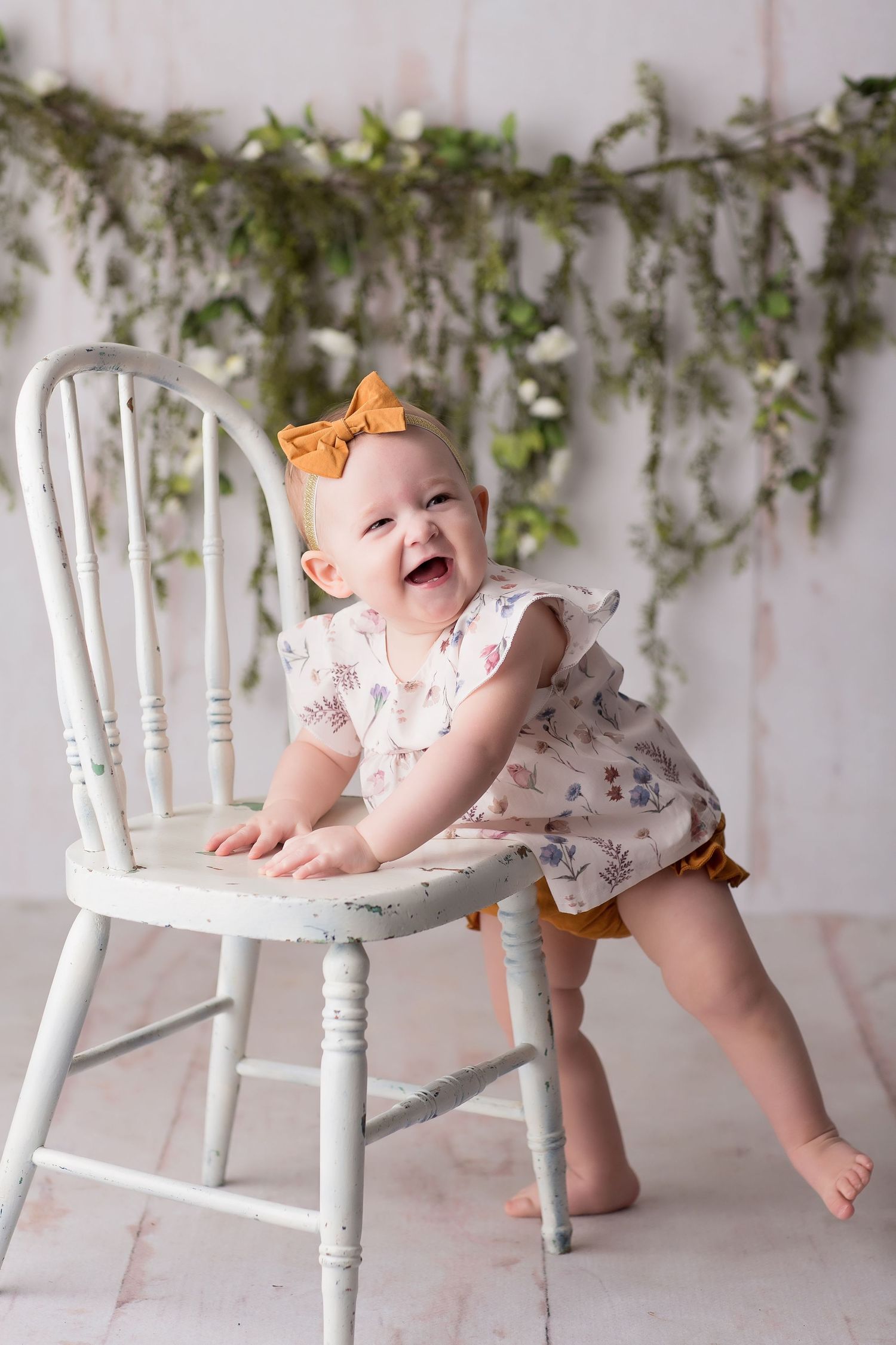 Baby in floral outfit with orange bow plays at white vintage chair under hanging greenery backdrop.