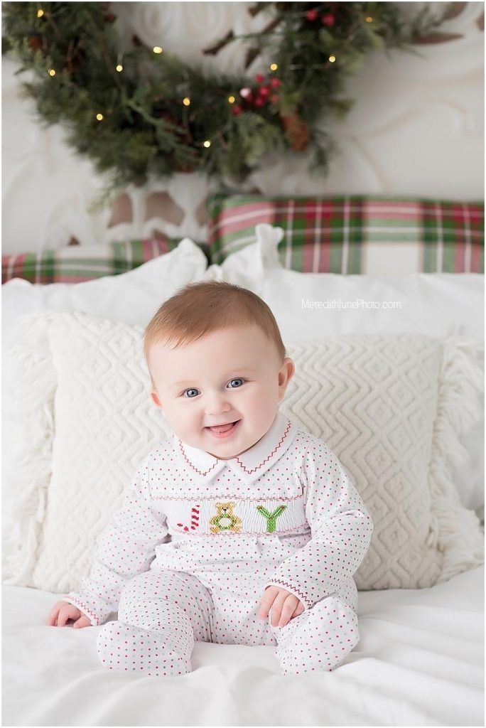 Baby wearing white polka dot holiday outfit smiles while lying on white bedding with Christmas decorations in background.
