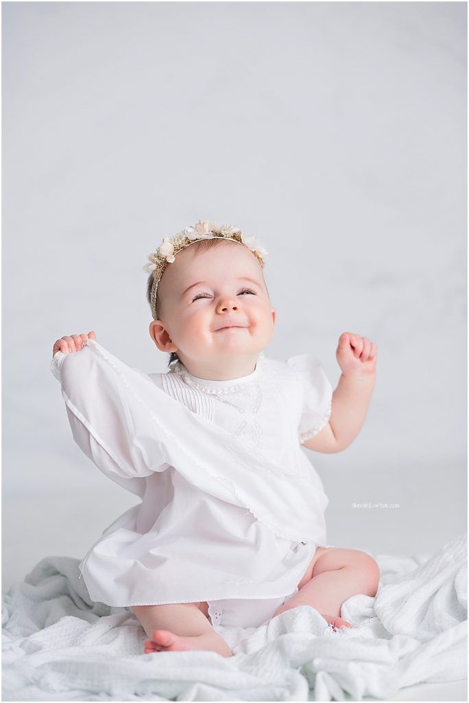 Baby wearing white dress sits on white fabric with arms raised and joyful expression.