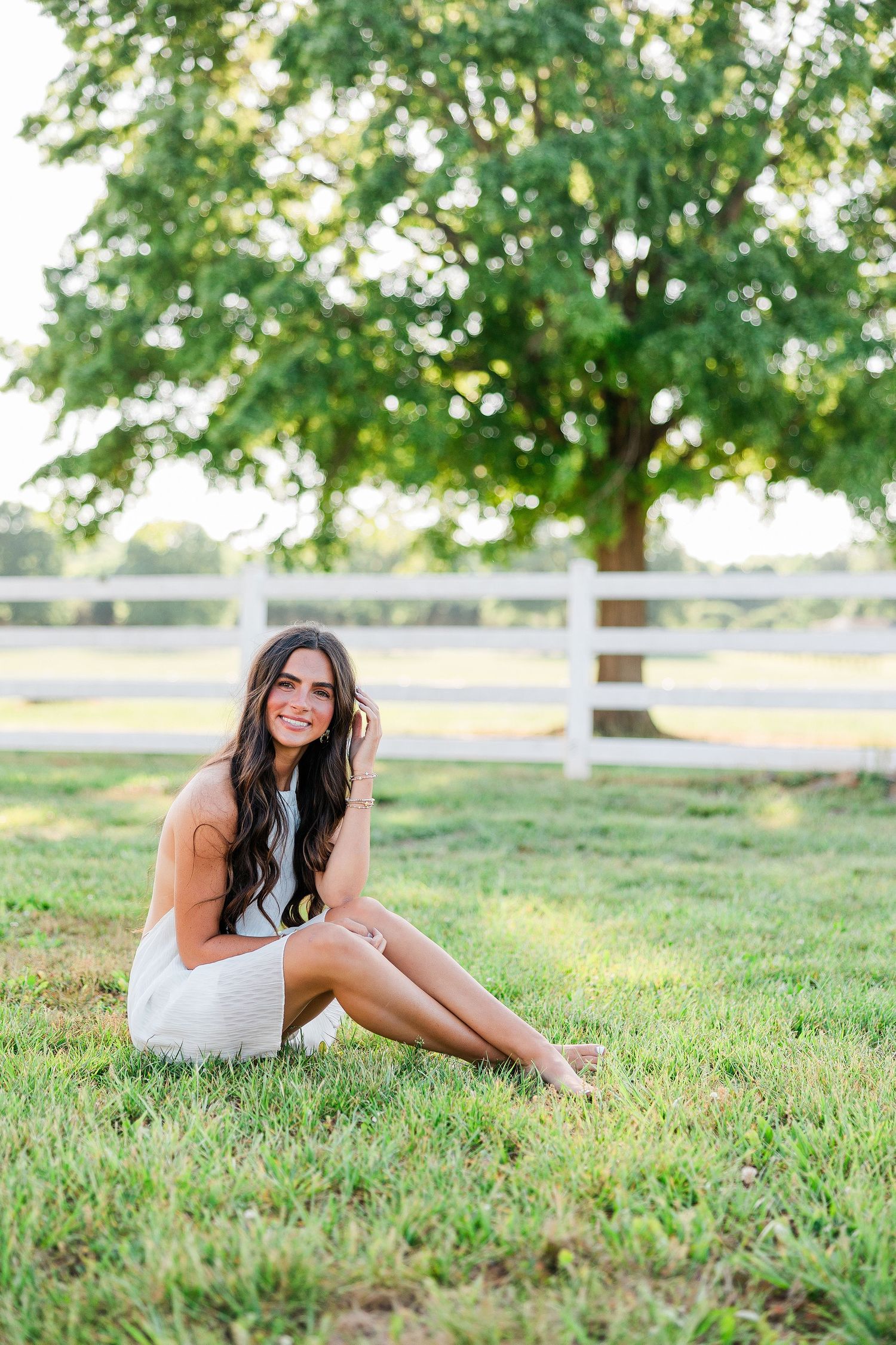 Smiling brunette woman in white mini dress sitting on green grass with white fence and tree backdrop.