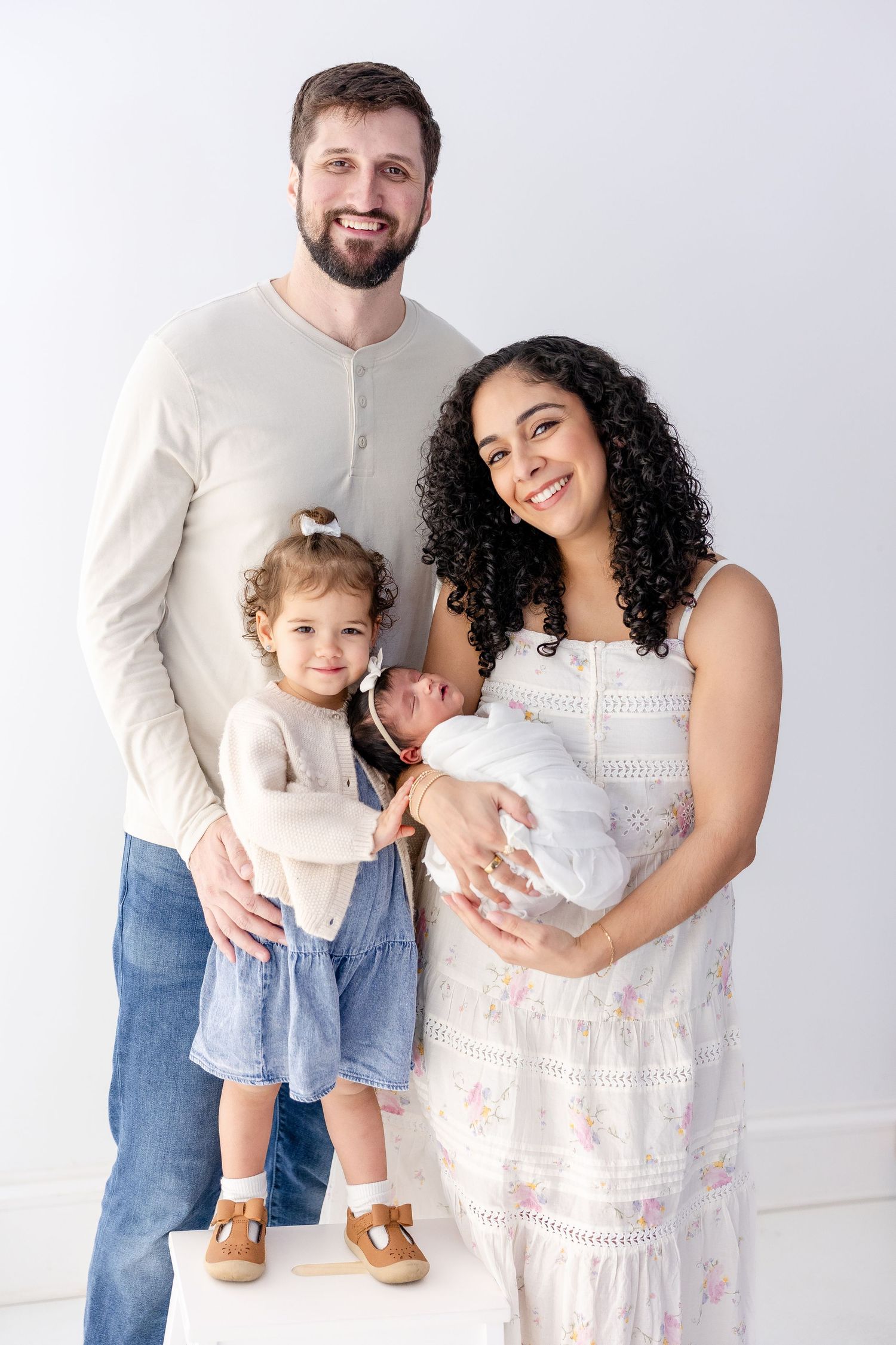 A family of four poses together against a white background wearing light colored clothing.