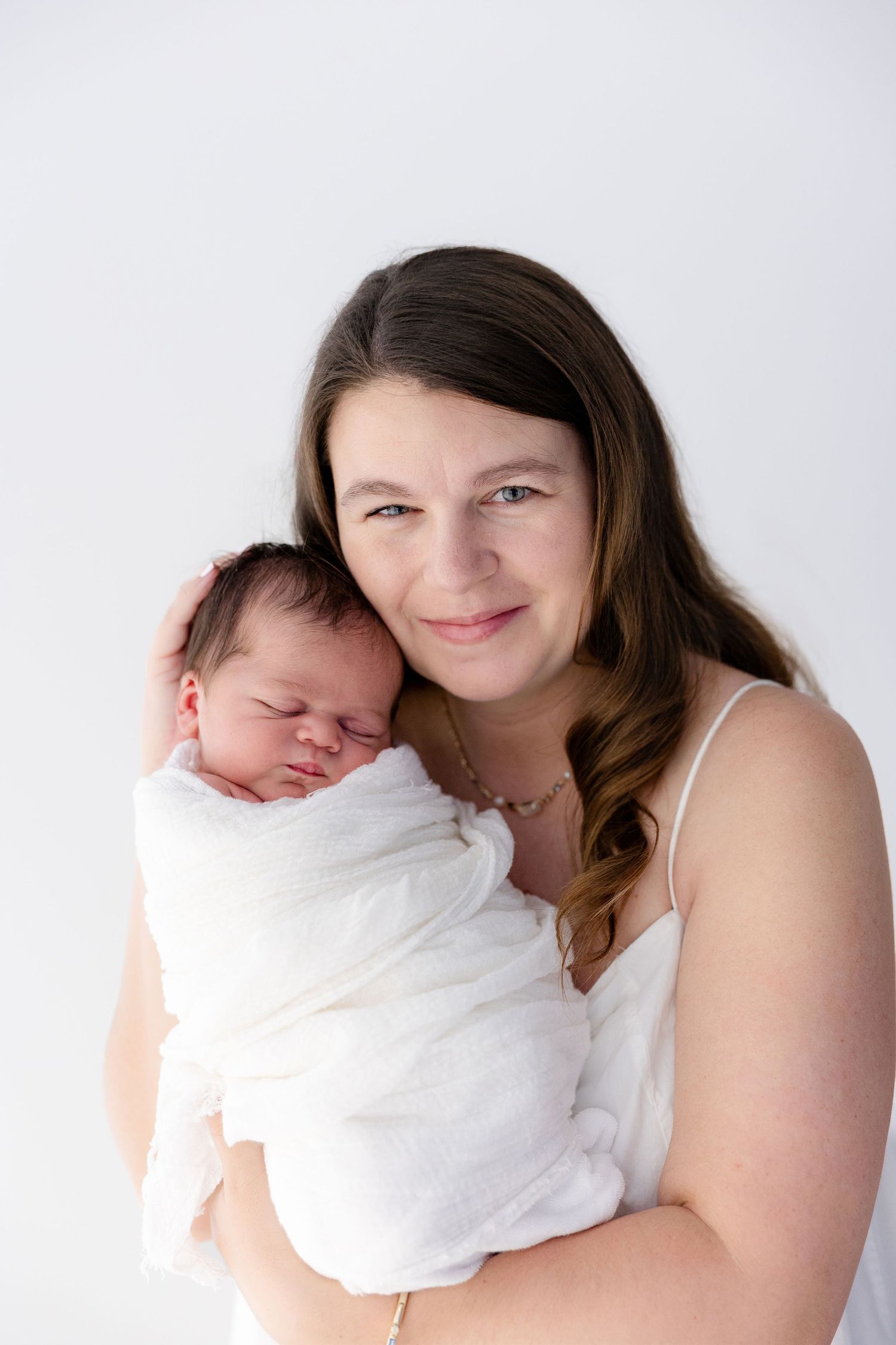 A smiling mother in a white dress holds her sleeping newborn baby wrapped in a white swaddle against a white background.