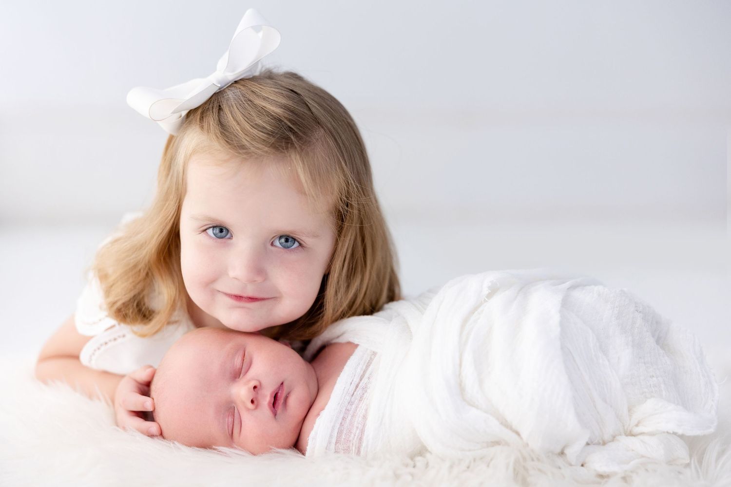 Young girl with blue eyes and white bow smiles beside swaddled newborn baby on white fluffy blanket.