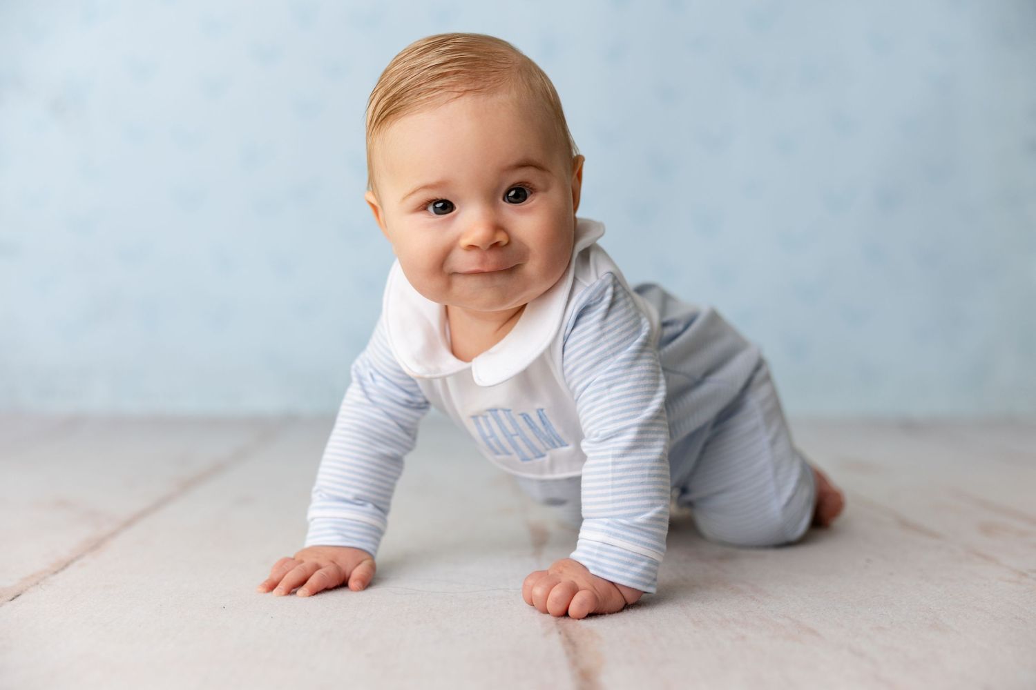 Baby in white onesie crawls and plays on light wooden floor against soft blue wall background.