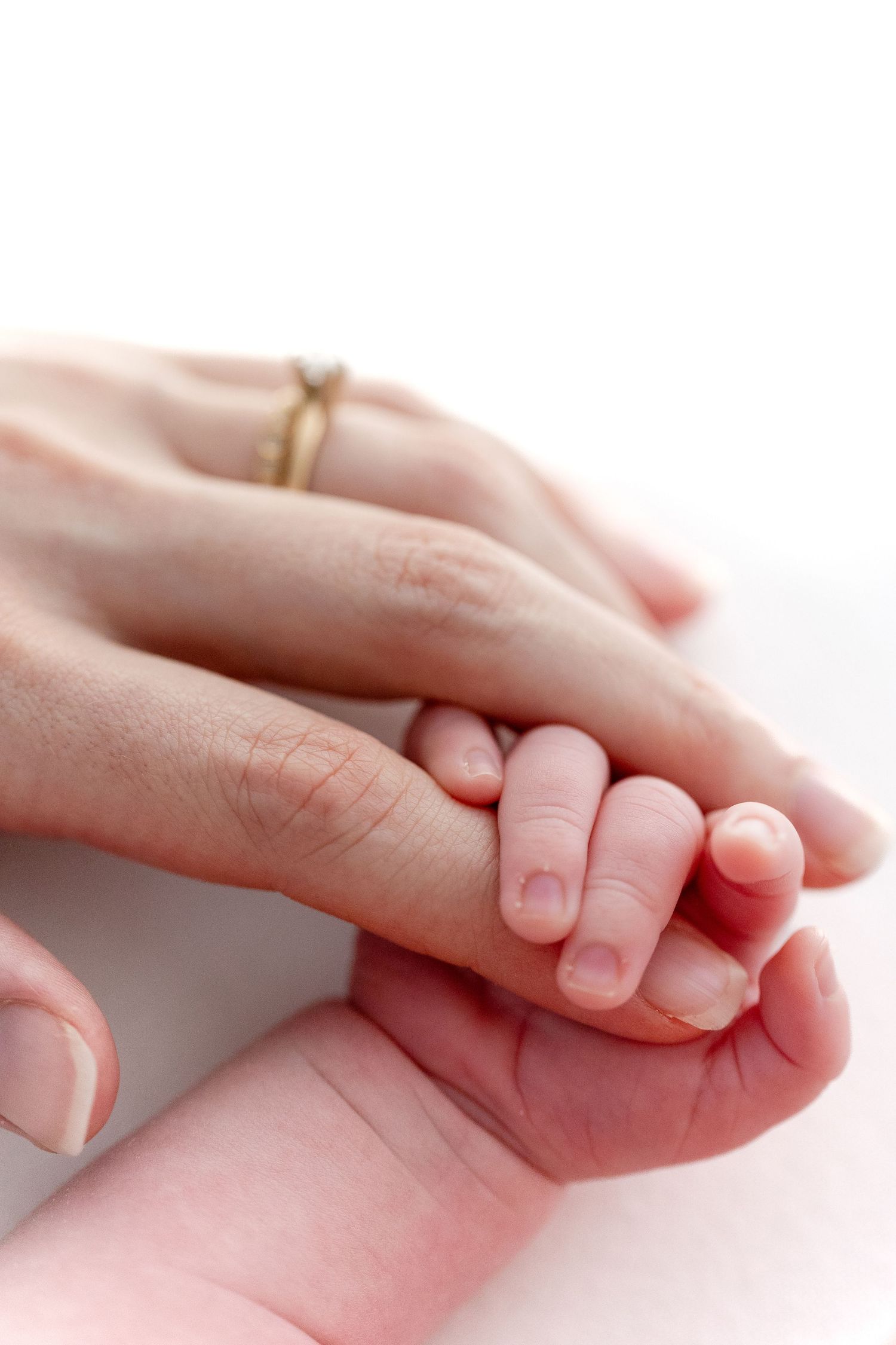 A mother's hand with a gold ring gently holds her newborn baby's tiny hand in a tender, loving moment.