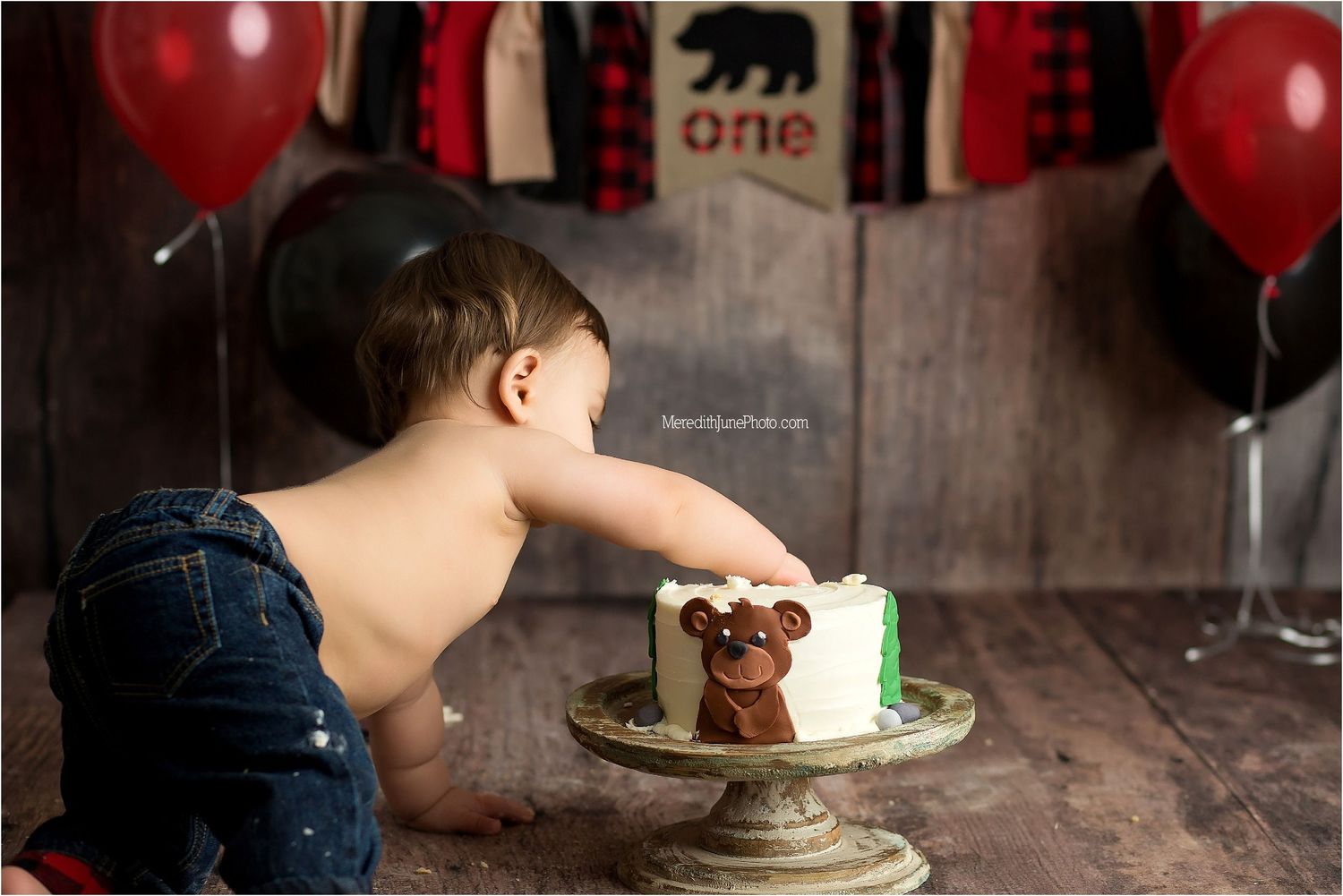 A birthday cake with a single candle is photographed in a rustic setting with red balloons in the background.