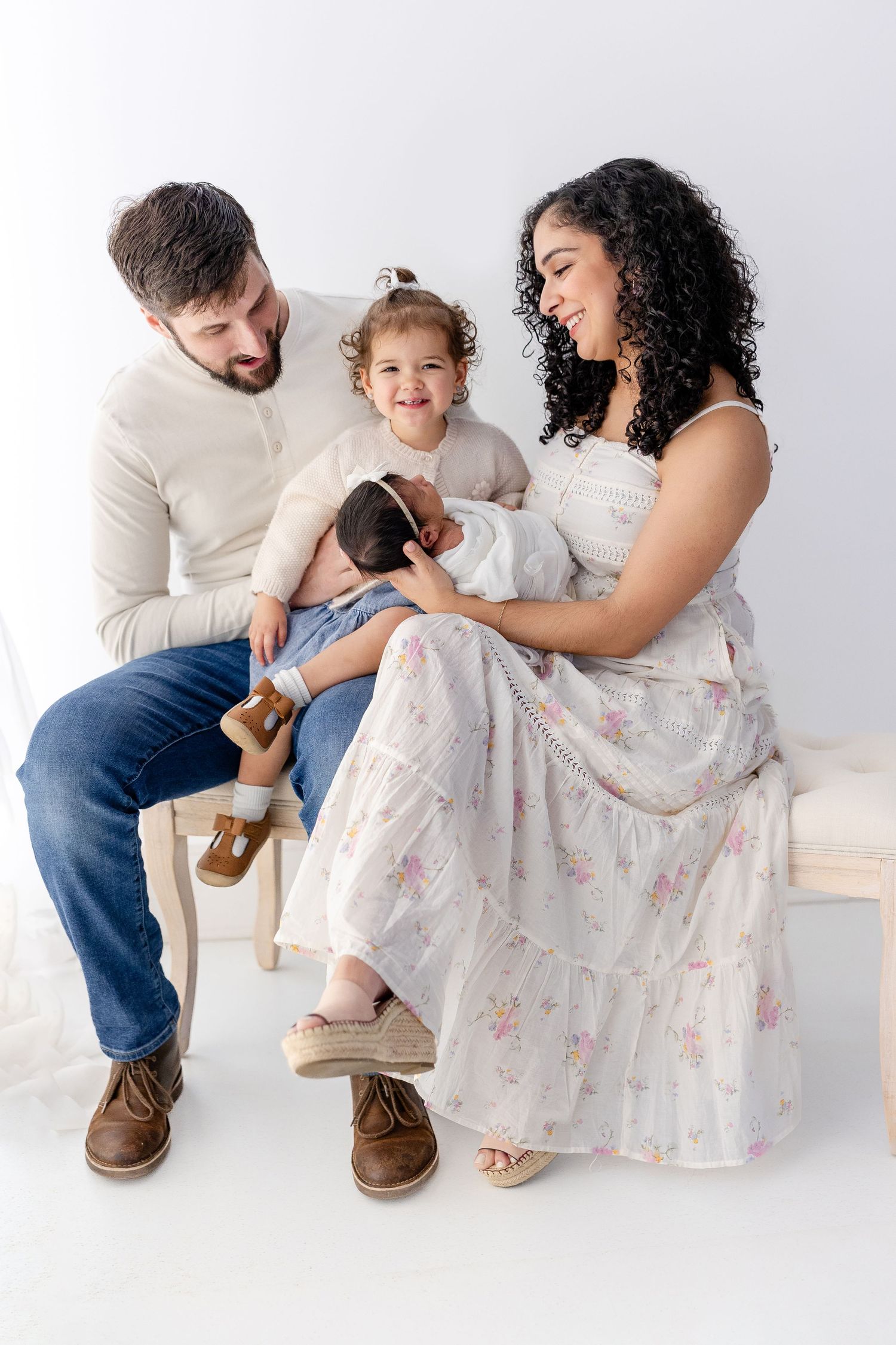 A family of three sits together on a white floor wearing coordinated cream and floral outfits in a minimalist studio setting.