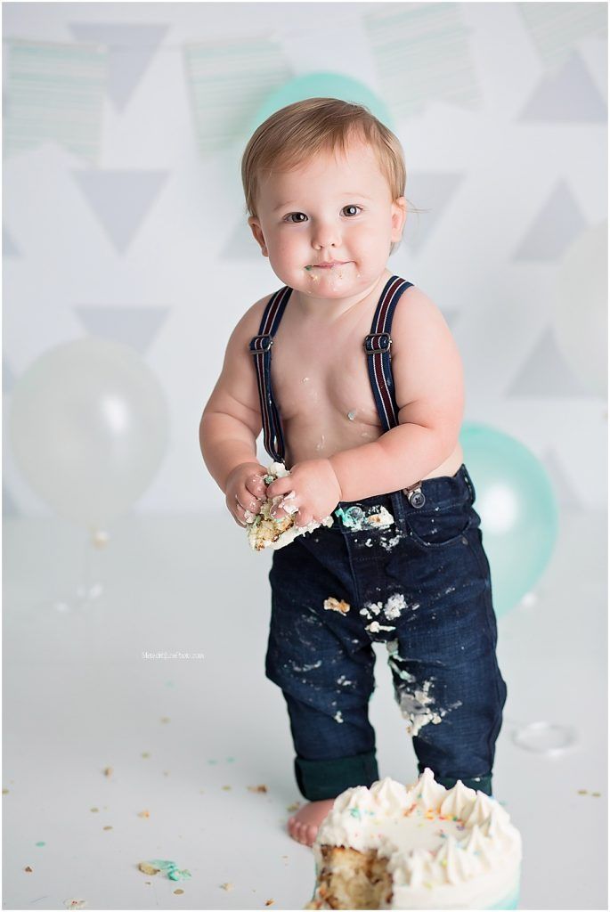 Baby in overalls enjoying a messy cake smash photo session with white frosting and teal decorations.