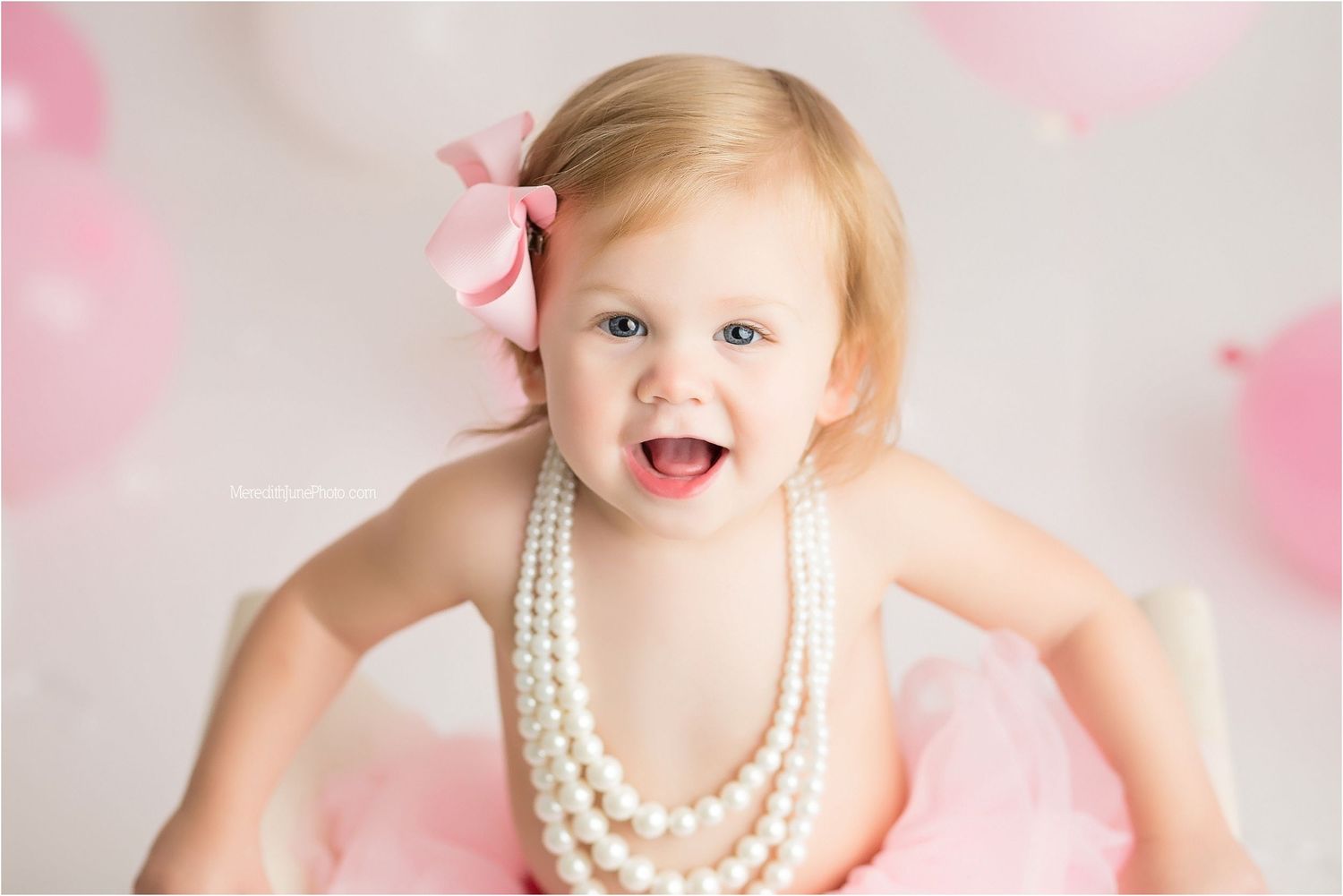 Baby wearing pearls and tutu smiles while surrounded by pink and white balloons for birthday photos.
