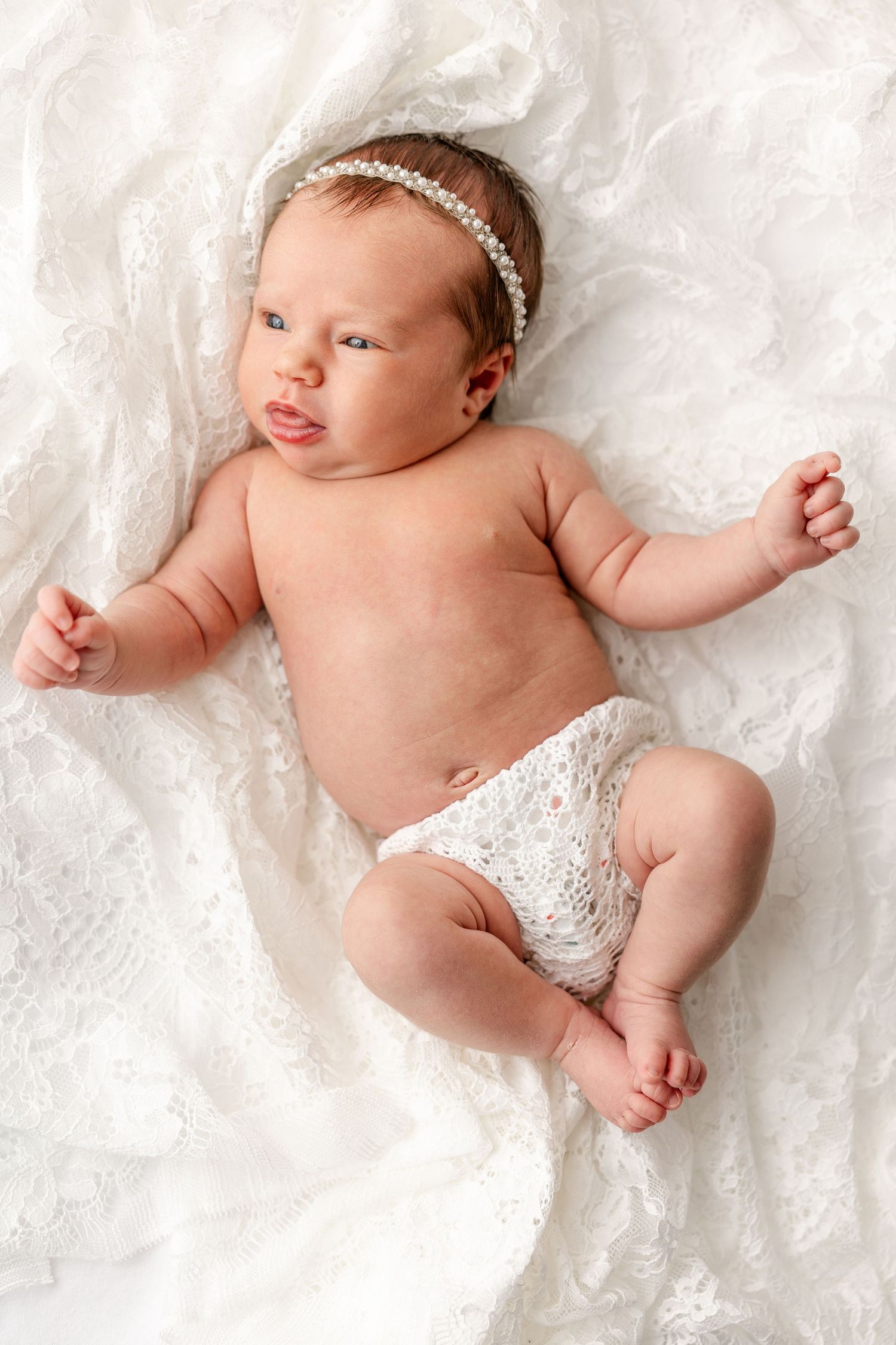 Newborn baby wearing a delicate headband and lace diaper lying on soft white fabric.