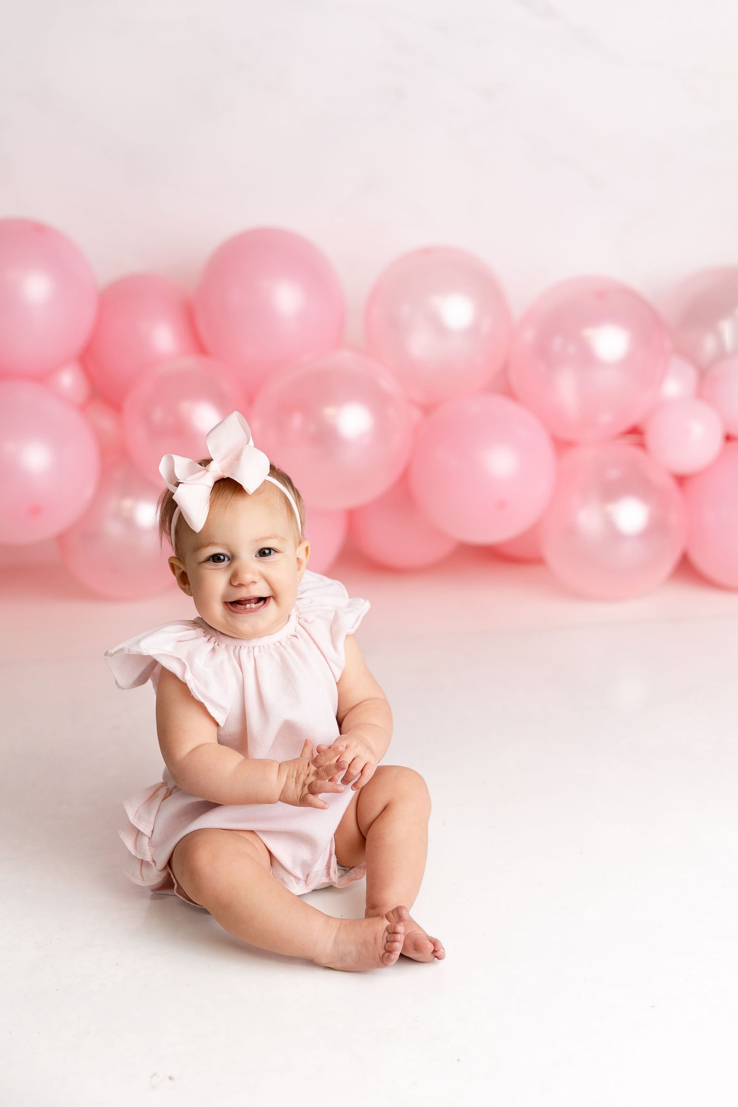 Baby in white outfit sits and plays against pink balloon backdrop during first birthday photoshoot.