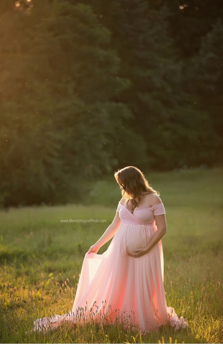 A pregnant woman in a pink flowing dress sits in a sunlit meadow during golden hour.