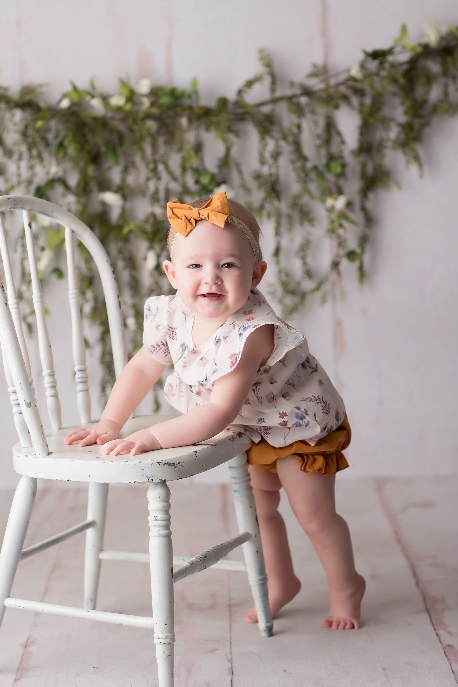 A baby in a white dress and orange bow sits playfully on white wooden chairs against a leafy backdrop.