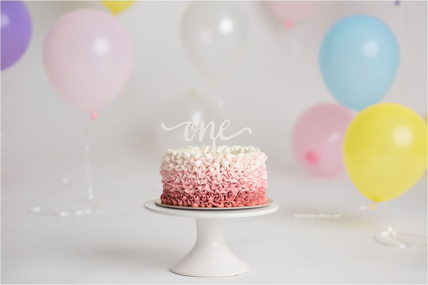 Pink and white sprinkled birthday cake on pedestal with colorful balloons in soft pastel background.