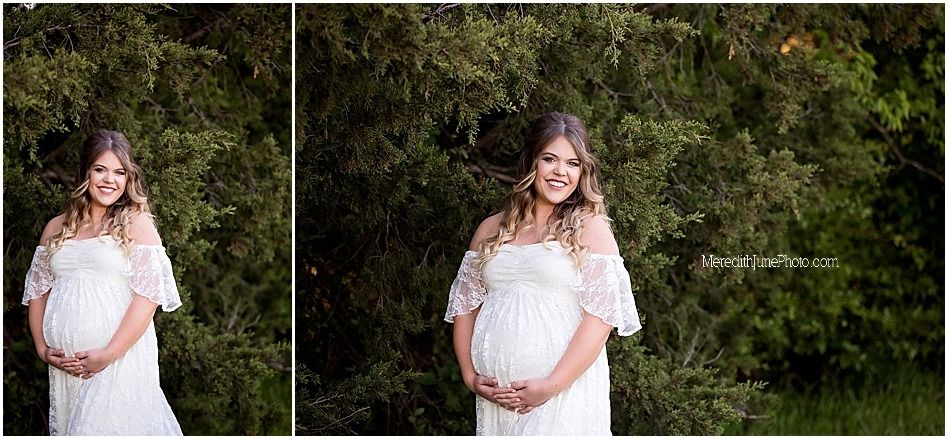 A woman in a white lace maternity dress poses gracefully in a lush garden setting during a photo session.