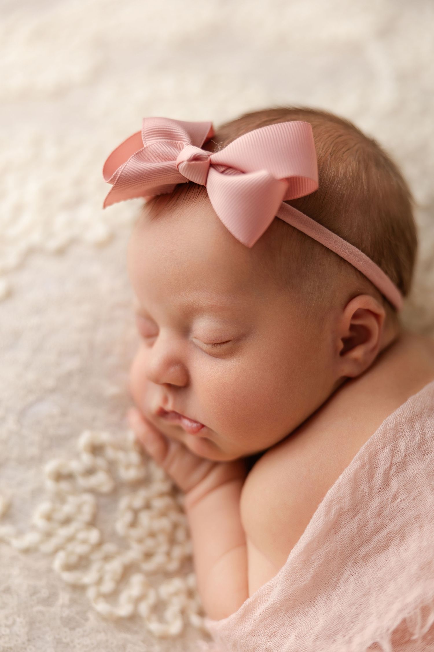 Sleeping newborn wearing pink bow headband rests peacefully on cream lace blanket.