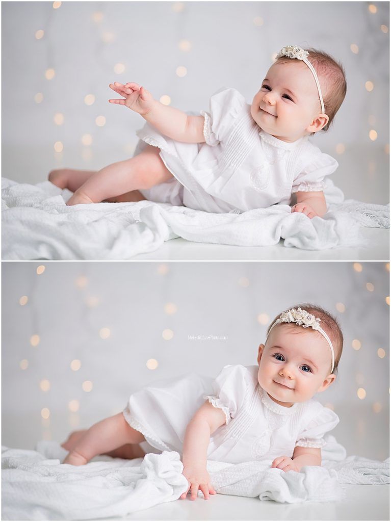 Sweet baby in white lace dress and headband smiles while sitting on soft white blanket with bokeh lights in background.