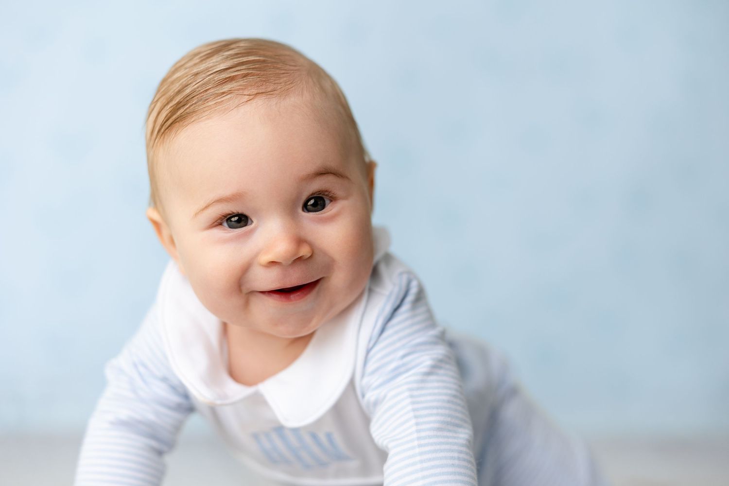 A smiling baby in a white striped outfit lies on their stomach against a light blue background.