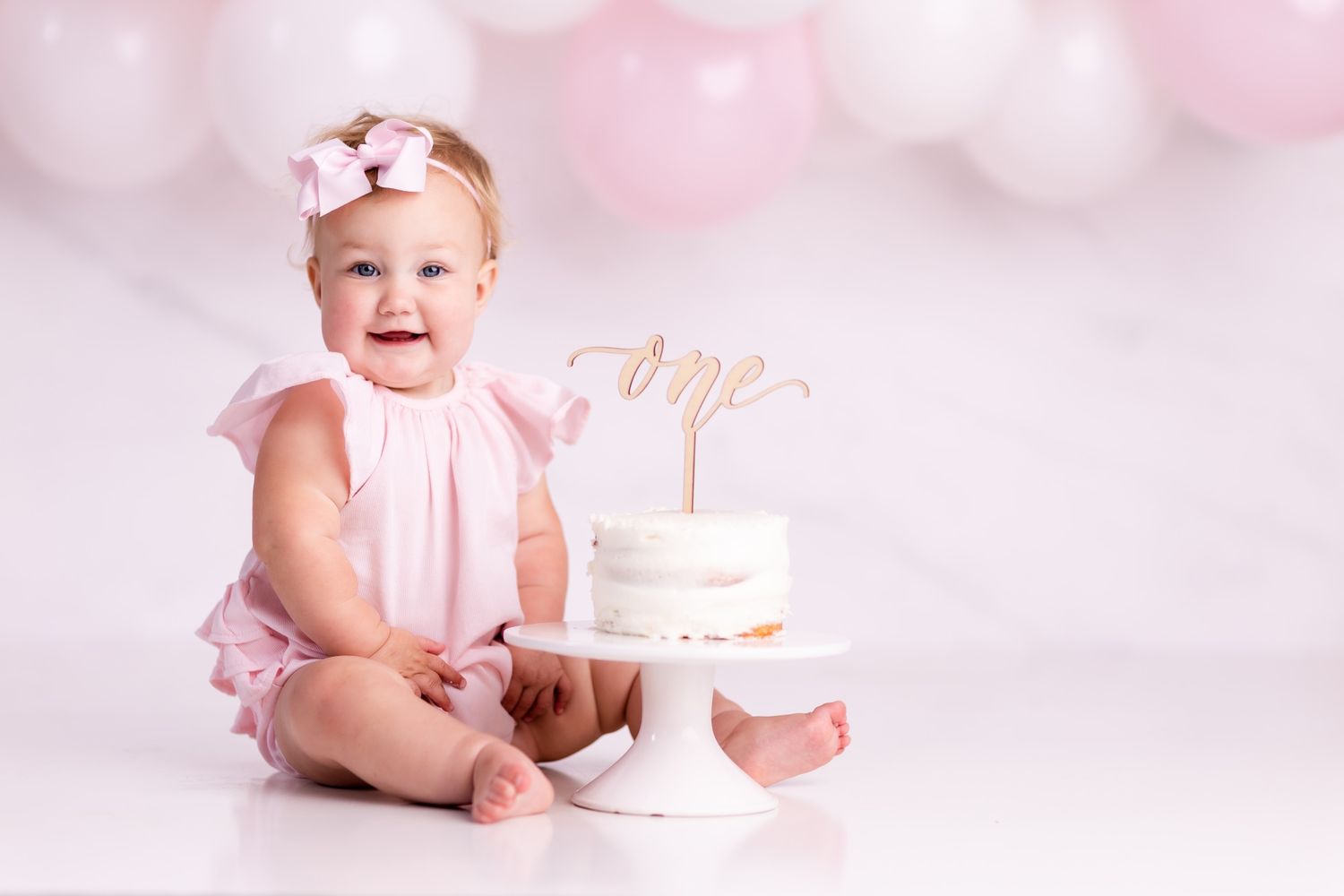 Baby in pink dress celebrating first birthday with cake on pink background.