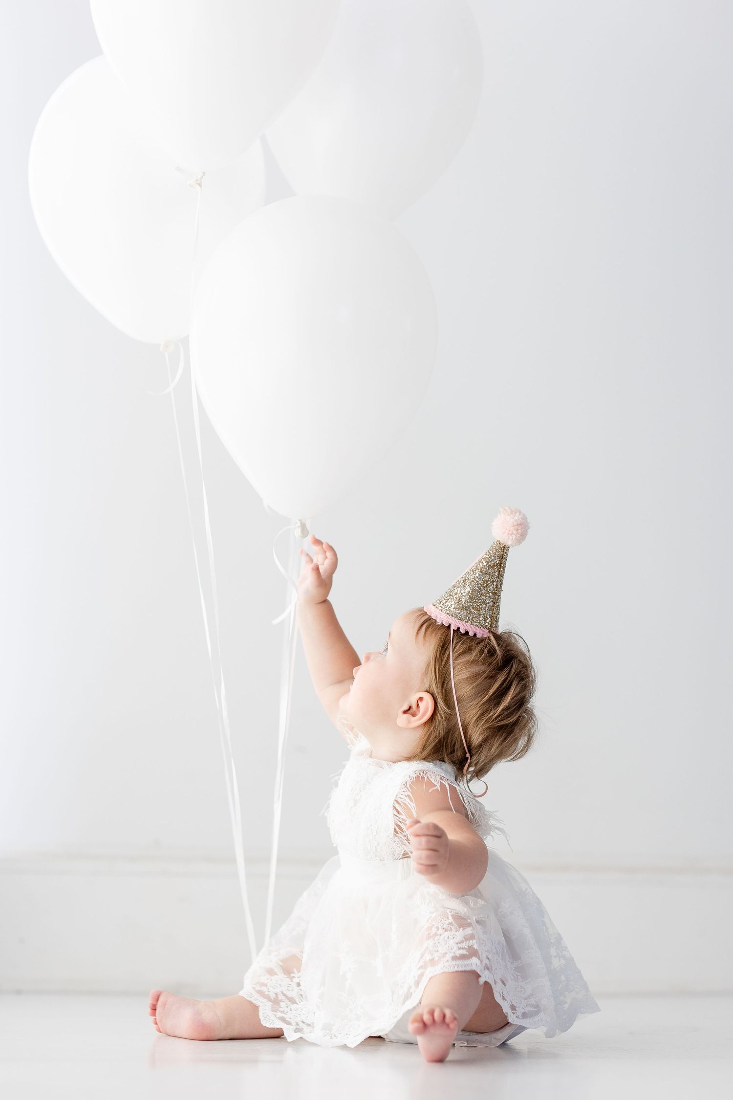 Baby in white dress plays with balloons during studio photography session.