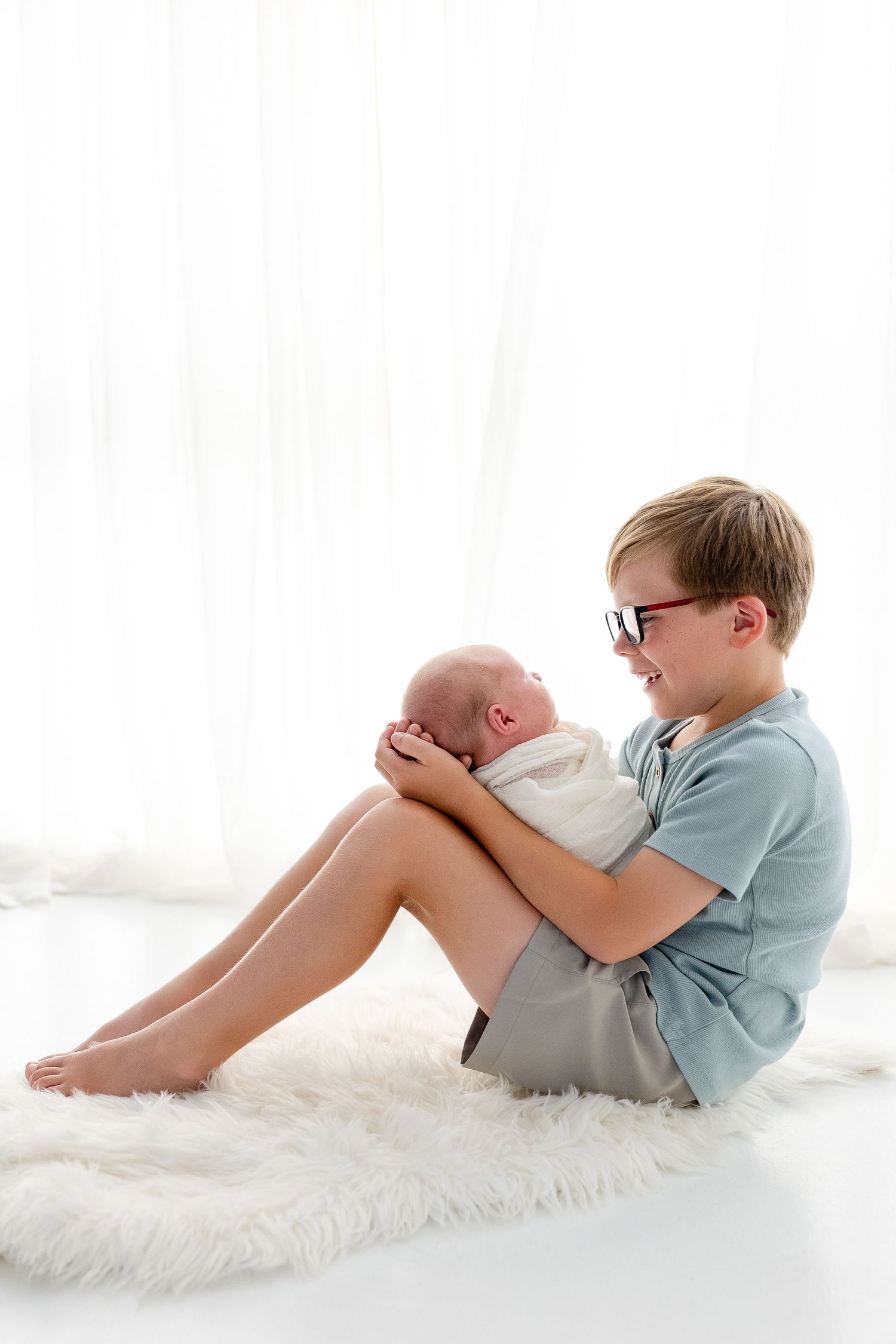 A smiling boy with glasses sits on a fluffy white rug, gently cradling a swaddled newborn baby.