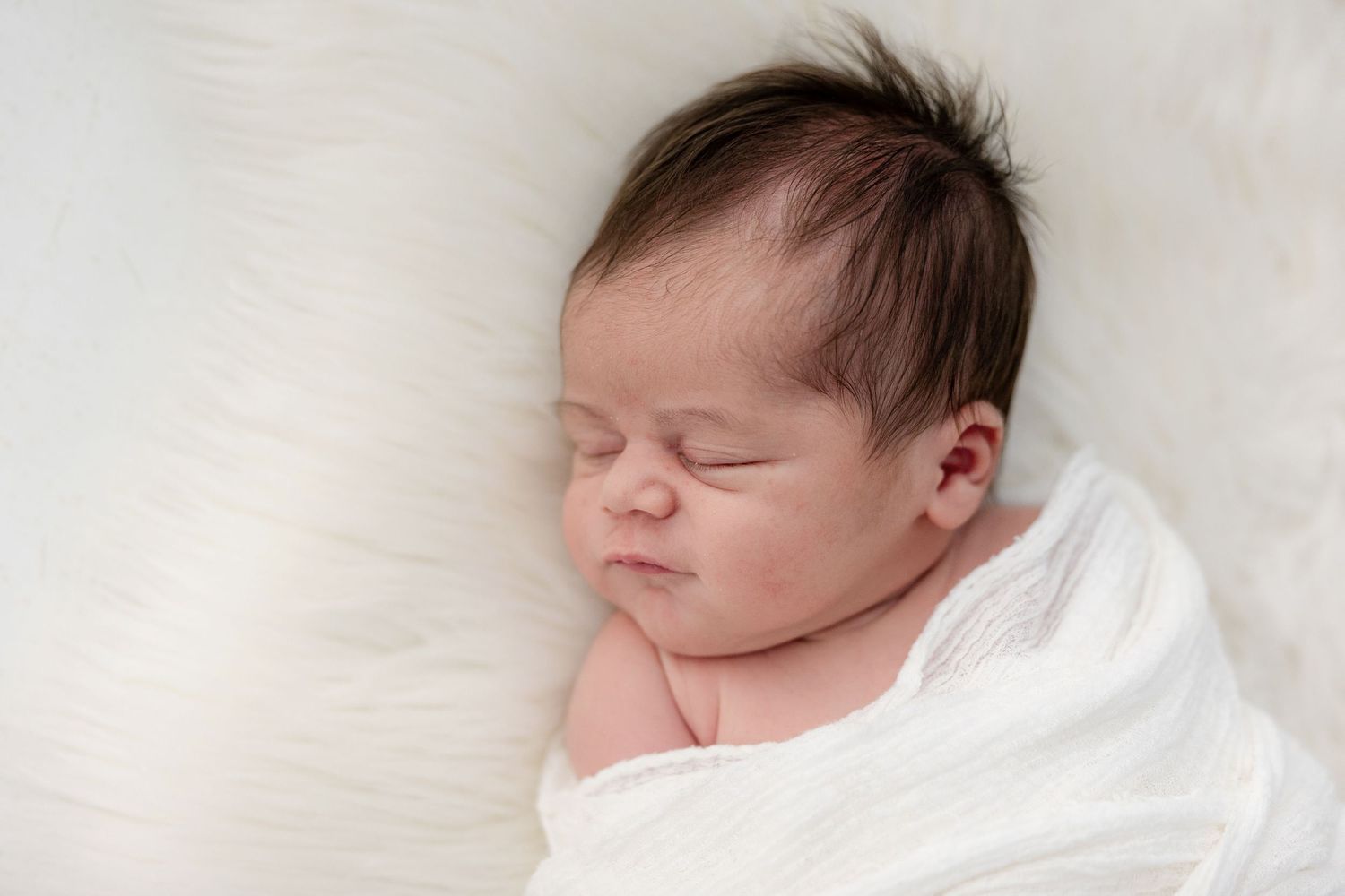 Peaceful newborn baby swaddled in white wrap sleeping on soft fluffy white fur blanket.