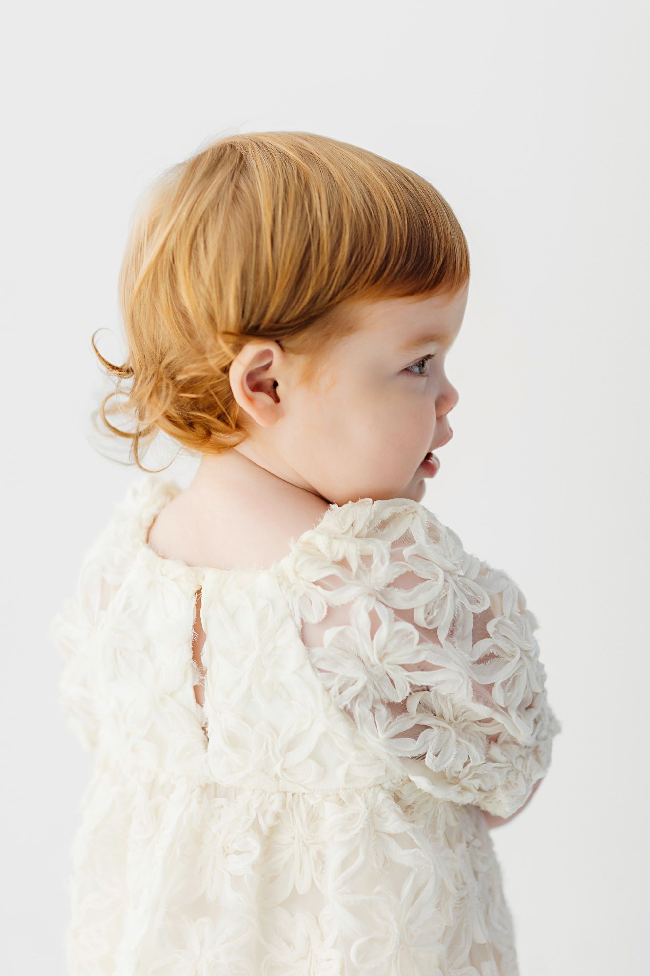 Side profile of toddler wearing delicate white lace dress against white background.