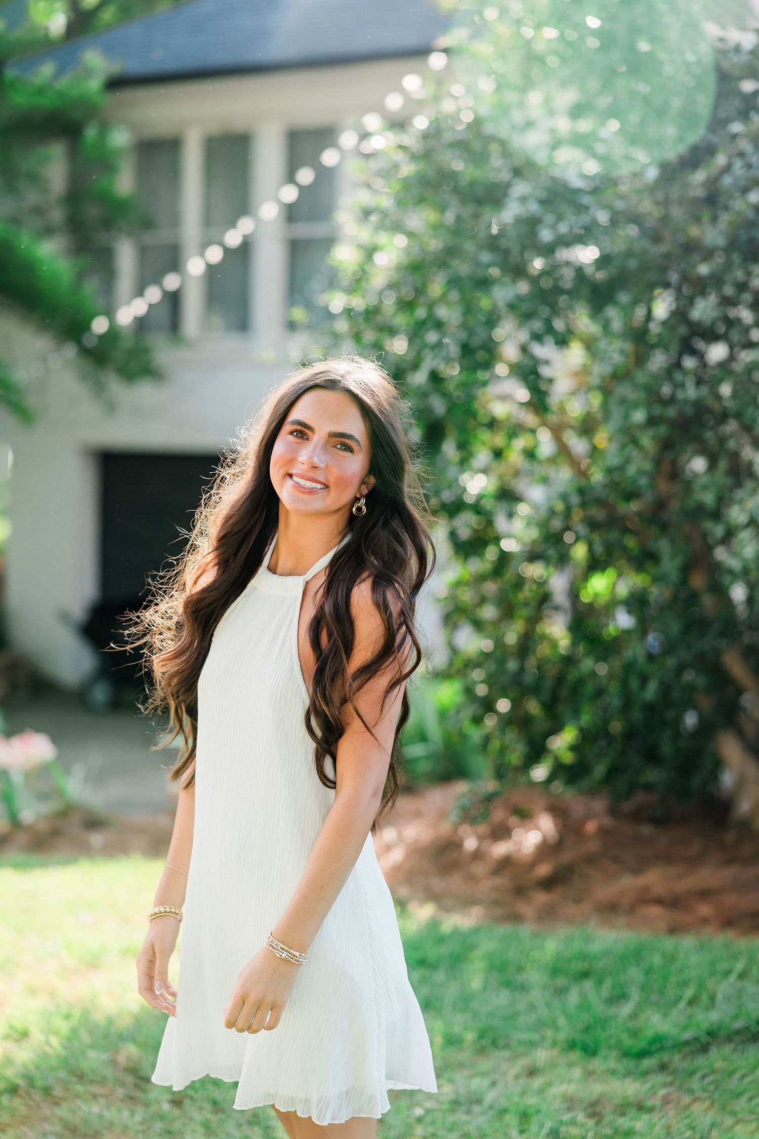 Smiling young woman with long dark hair wearing a white dress standing in a sunny backyard garden.