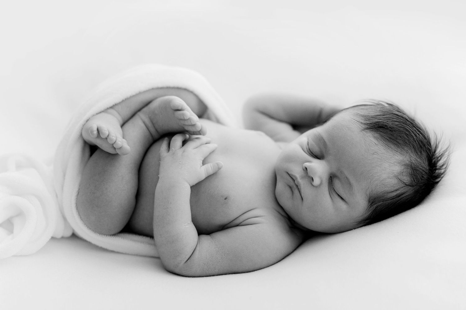 Black and white newborn baby sleeping peacefully curled up on white blanket, professional infant photography.