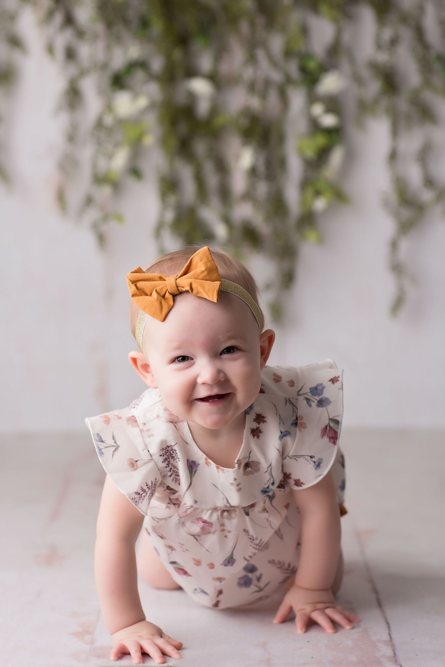 Baby in white floral dress with orange bow headband smiles while crawling on white floor.