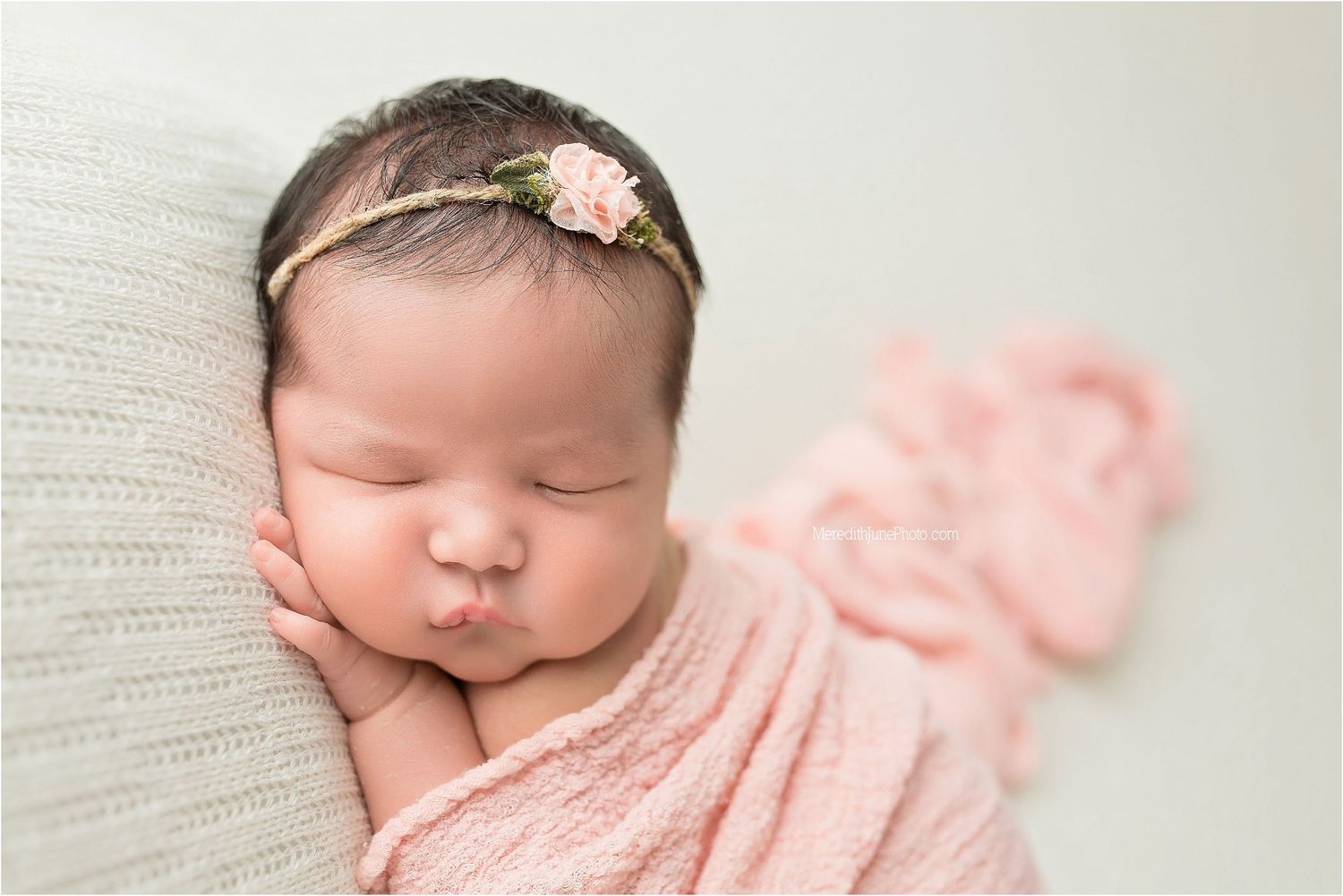 Sleeping newborn baby wrapped in soft pink blanket with delicate flower headband resting on white textured surface.