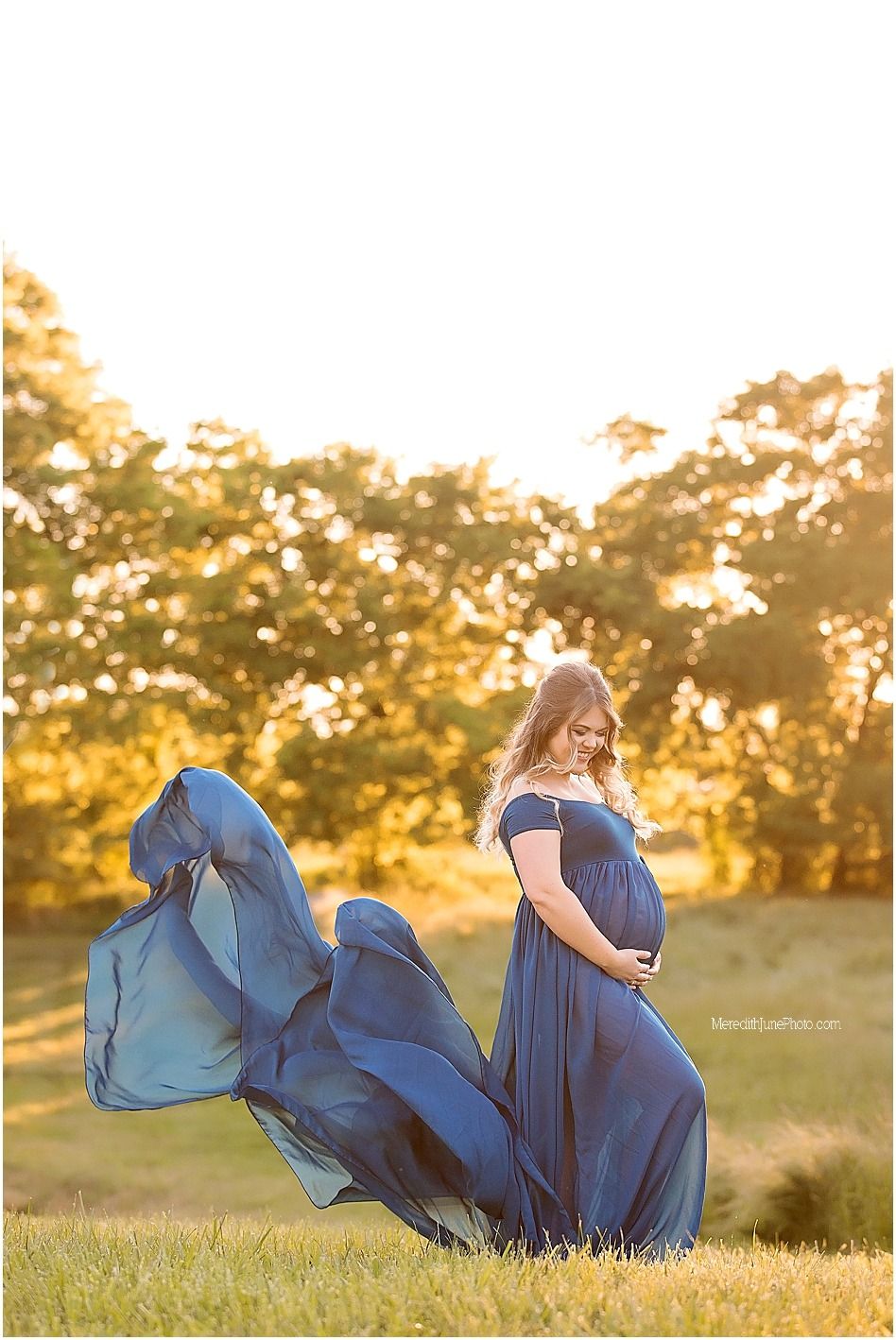 A flowing navy blue dress billows dramatically in a golden sunset field creating an ethereal maternity photo.