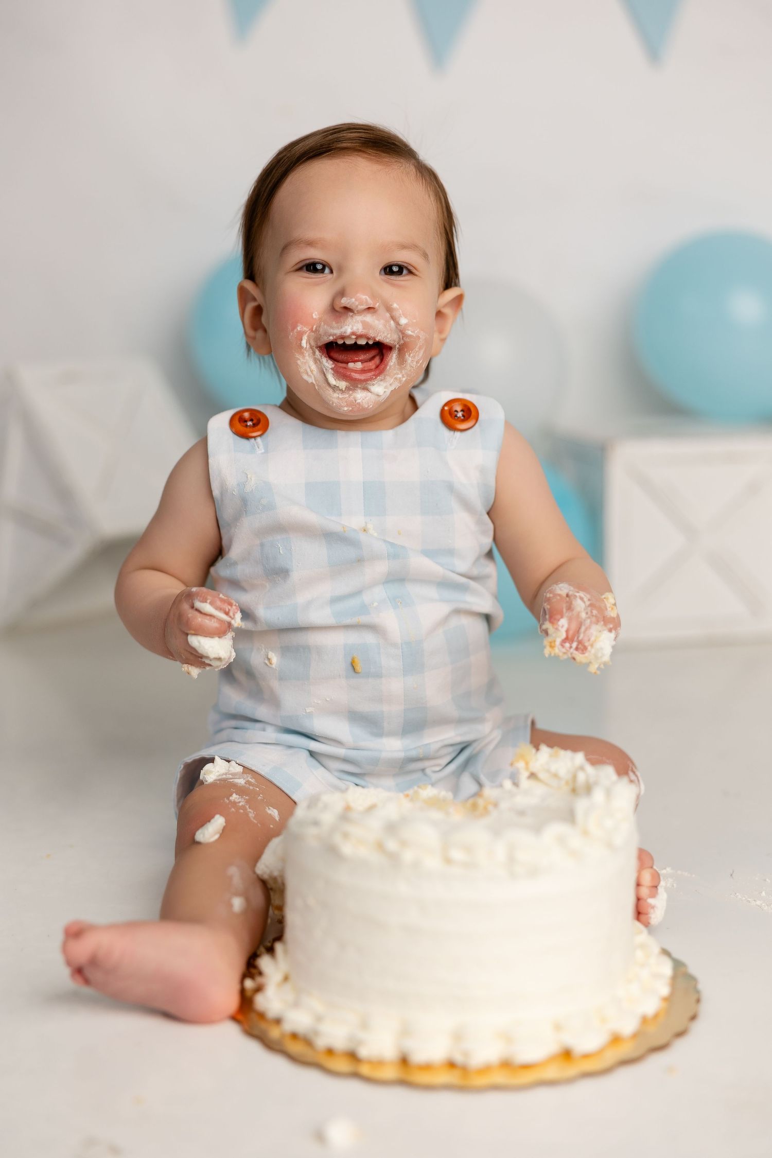 Baby in light blue gingham outfit laughs joyfully during cake smash photo session with white frosted cake.
