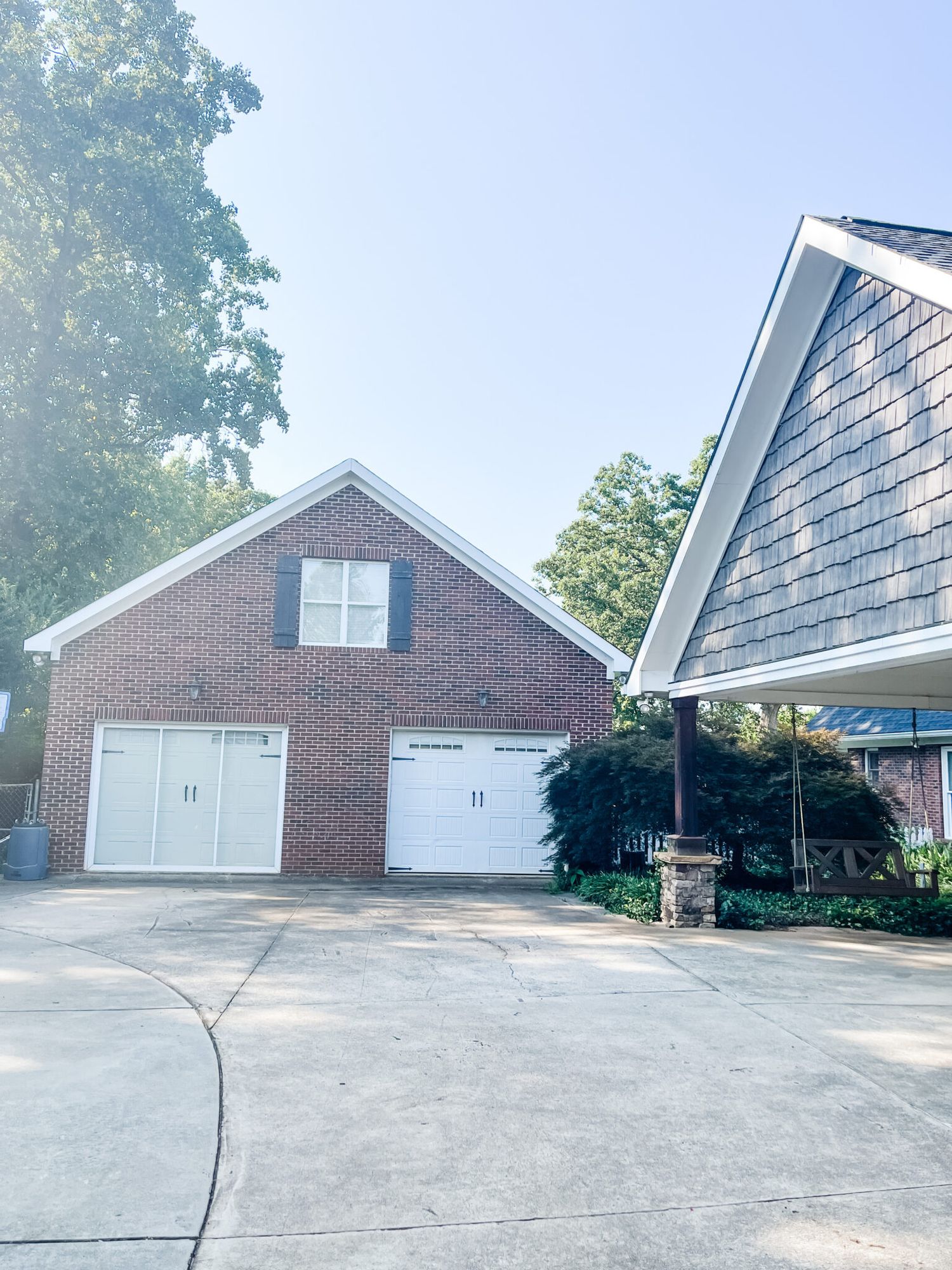 A brick detached garage with two white doors stands next to a house with gray siding and a covered porch.