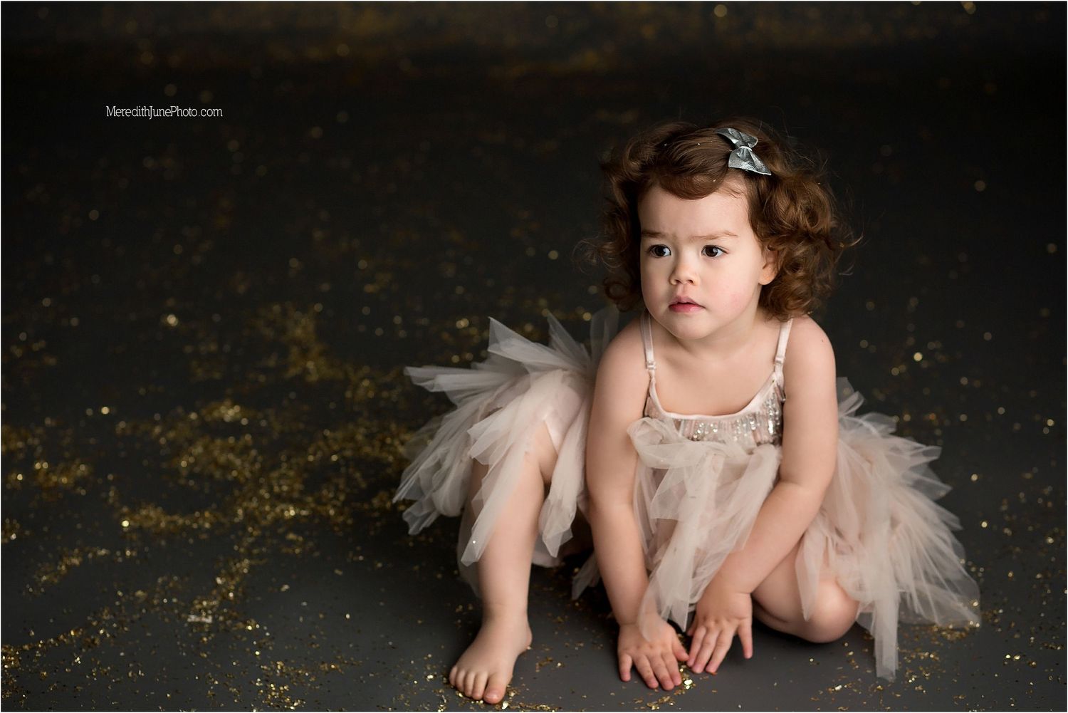A small child in a tulle dress poses on a dark floor with sparkly gold glitter scattered around.