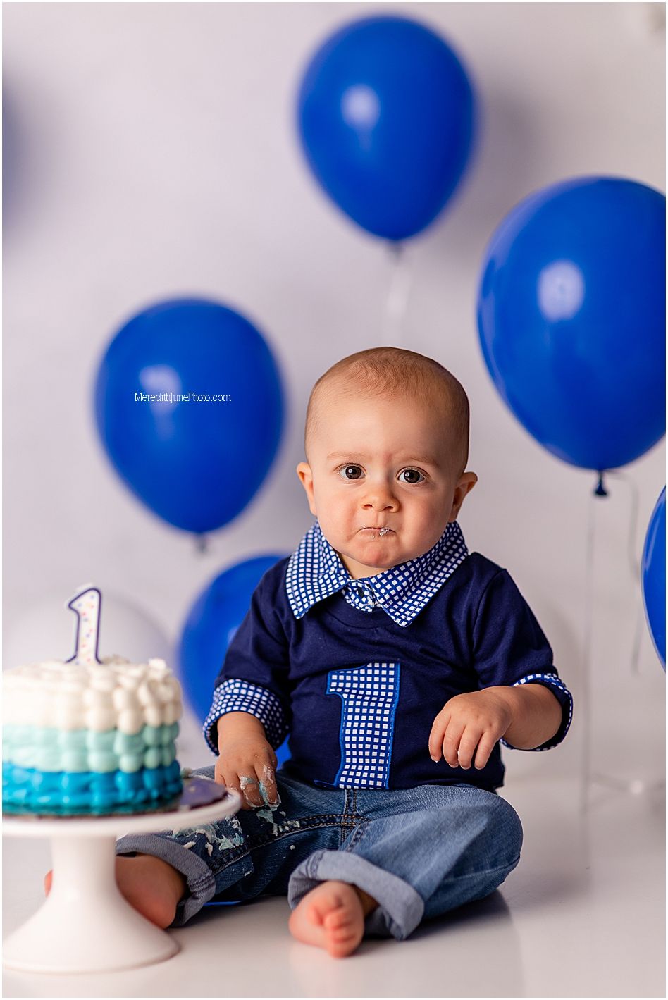 A baby's first birthday photoshoot with blue balloons, gingham shirt, and white cake against a gray backdrop.