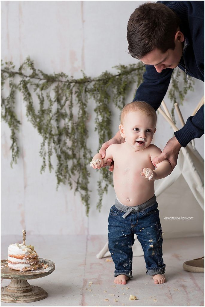 A baby in jeans sits on a white floor while being offered a slice of birthday cake during a cake smash photo session.