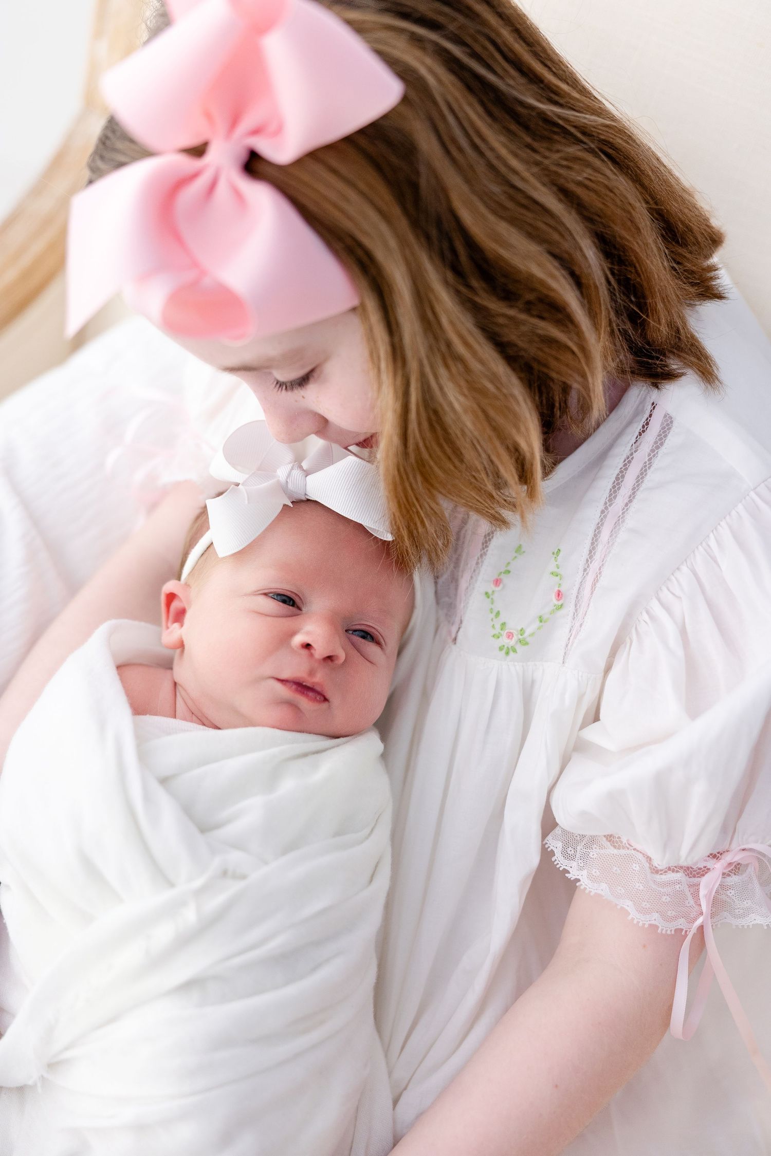 Sleeping baby wearing pink bow headband rests peacefully in white bedding with delicate floral details.