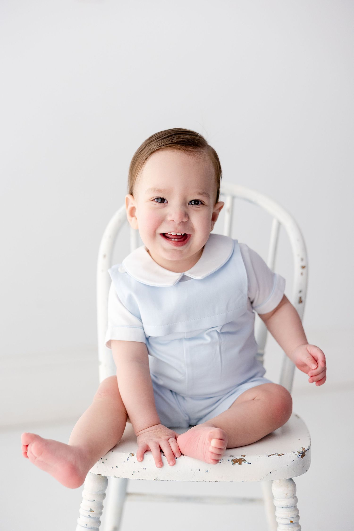 Happy baby in blue and white outfit poses on vintage white chair during studio photography session.