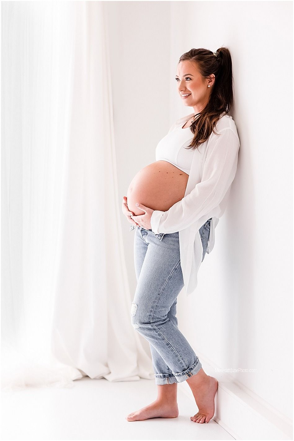 A minimalist maternity photo series showing a pregnant woman in jeans and white top against a white backdrop.