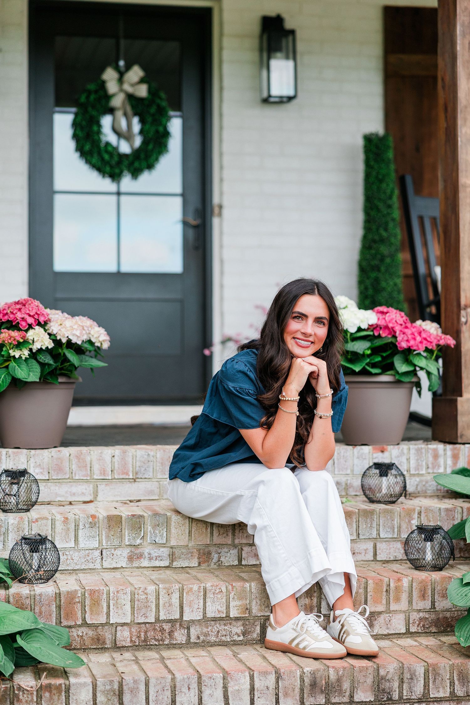 Woman in navy top and white pants sitting on brick porch steps with hydrangeas and wreath on door.