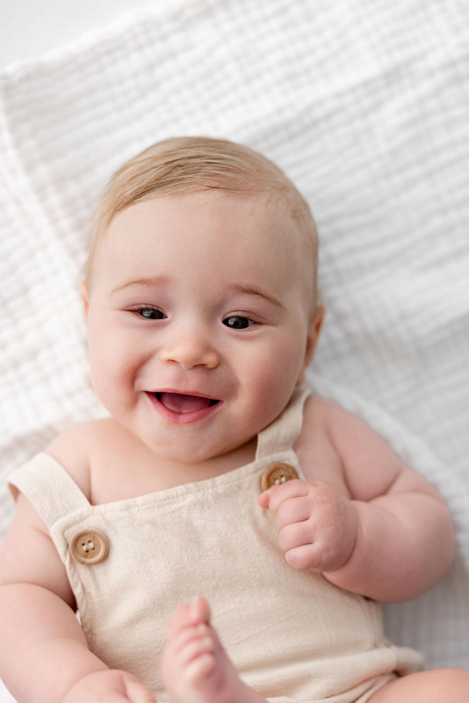 A smiling baby wearing a cream-colored romper lies on white bedding in a series of four connected photos.