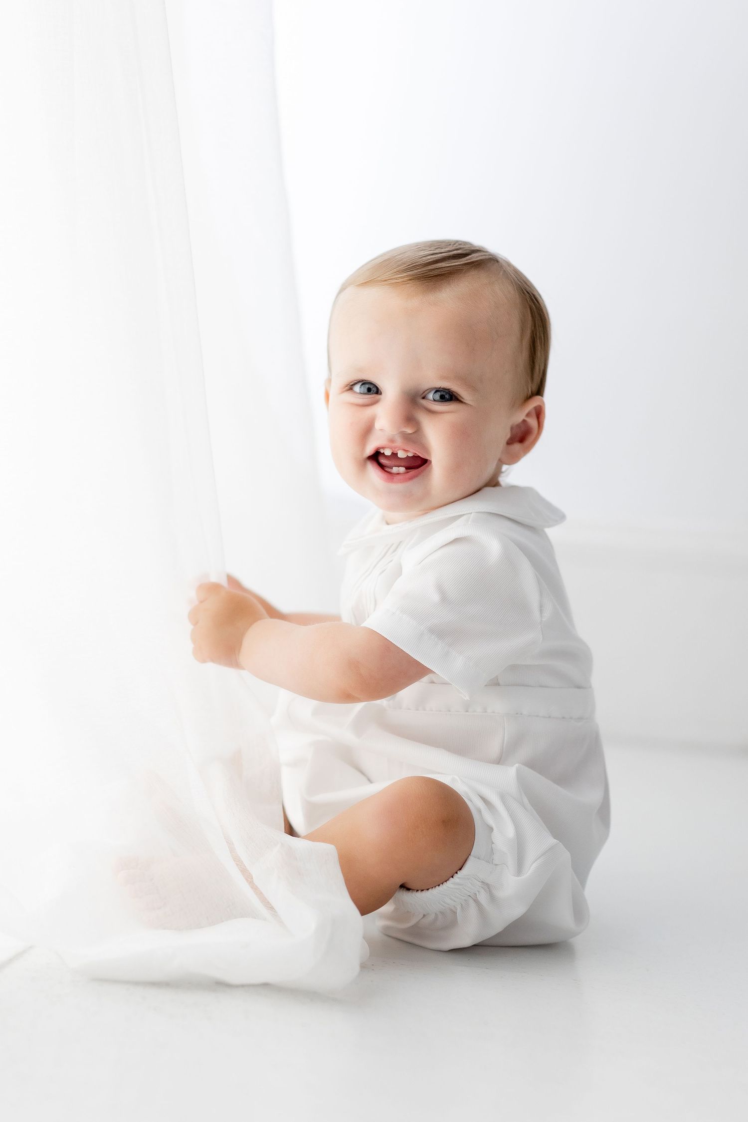 Baby in white romper sits and laughs during studio photoshoot.