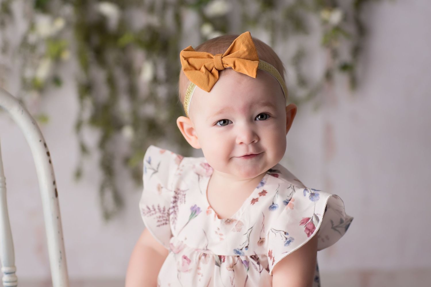Baby in ruffled white dress with orange bow sits on white floor with greenery backdrop.